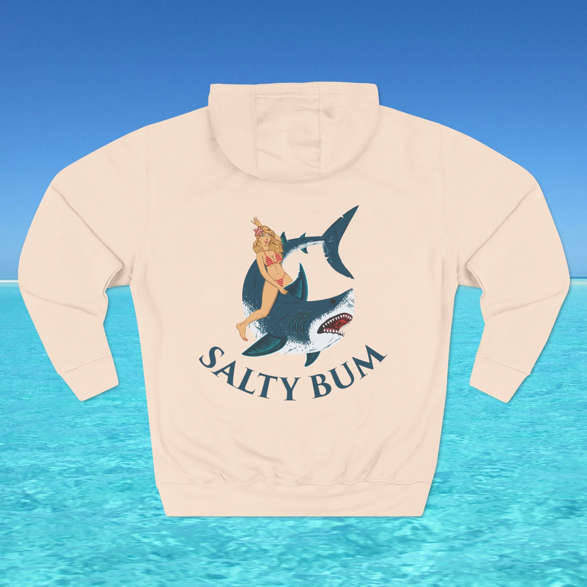Beige hoodie with a graphic of a woman on a shark and 'Salty Bum' text, set against a blue water and sky background.