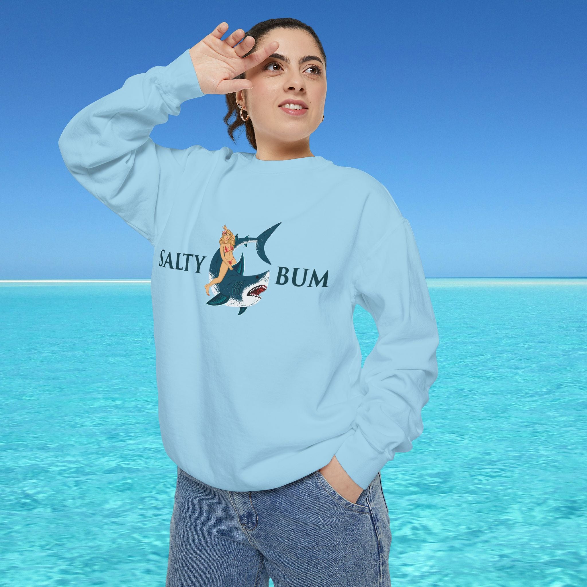 Person wearing a light blue sweatshirt with a graphic design and text, standing in front of a clear blue sky and ocean.
