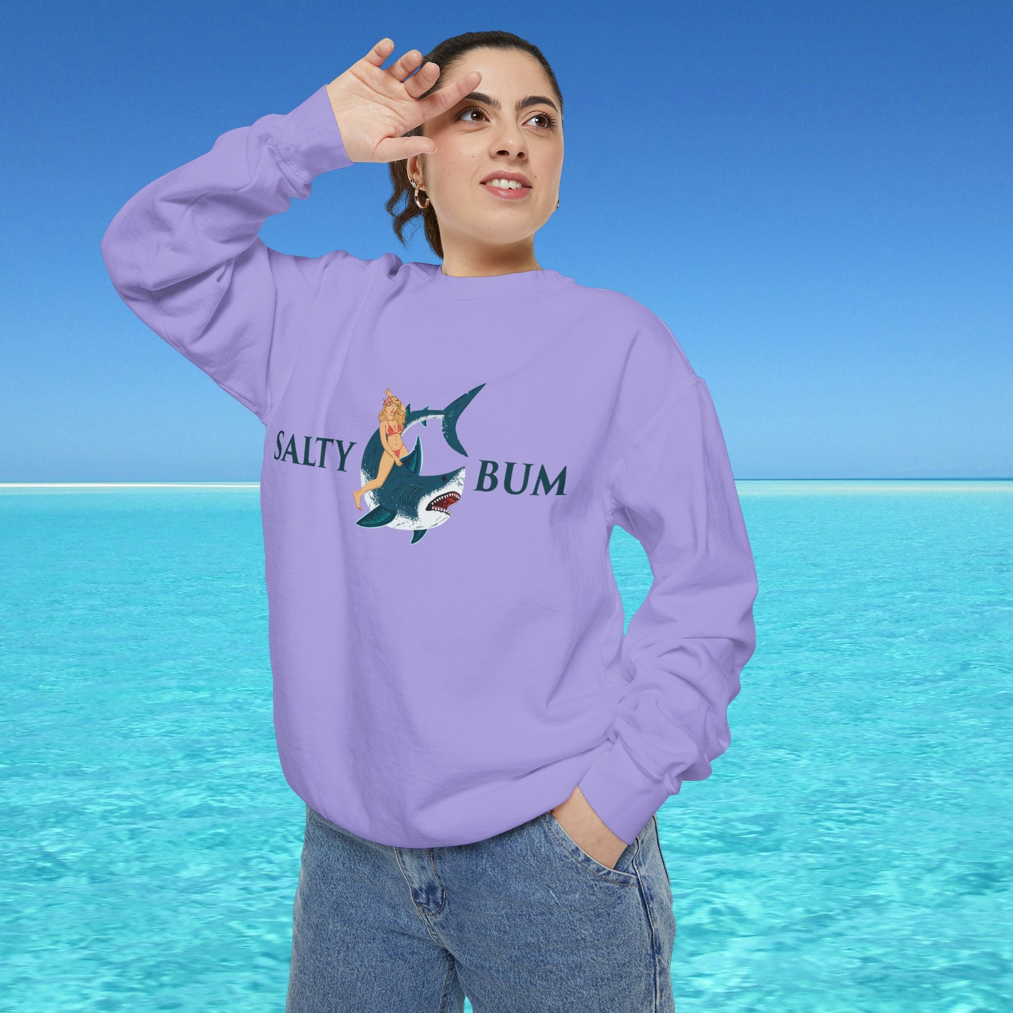 Person wearing a purple sweatshirt with a graphic design and text, standing against a blue sky and ocean background.