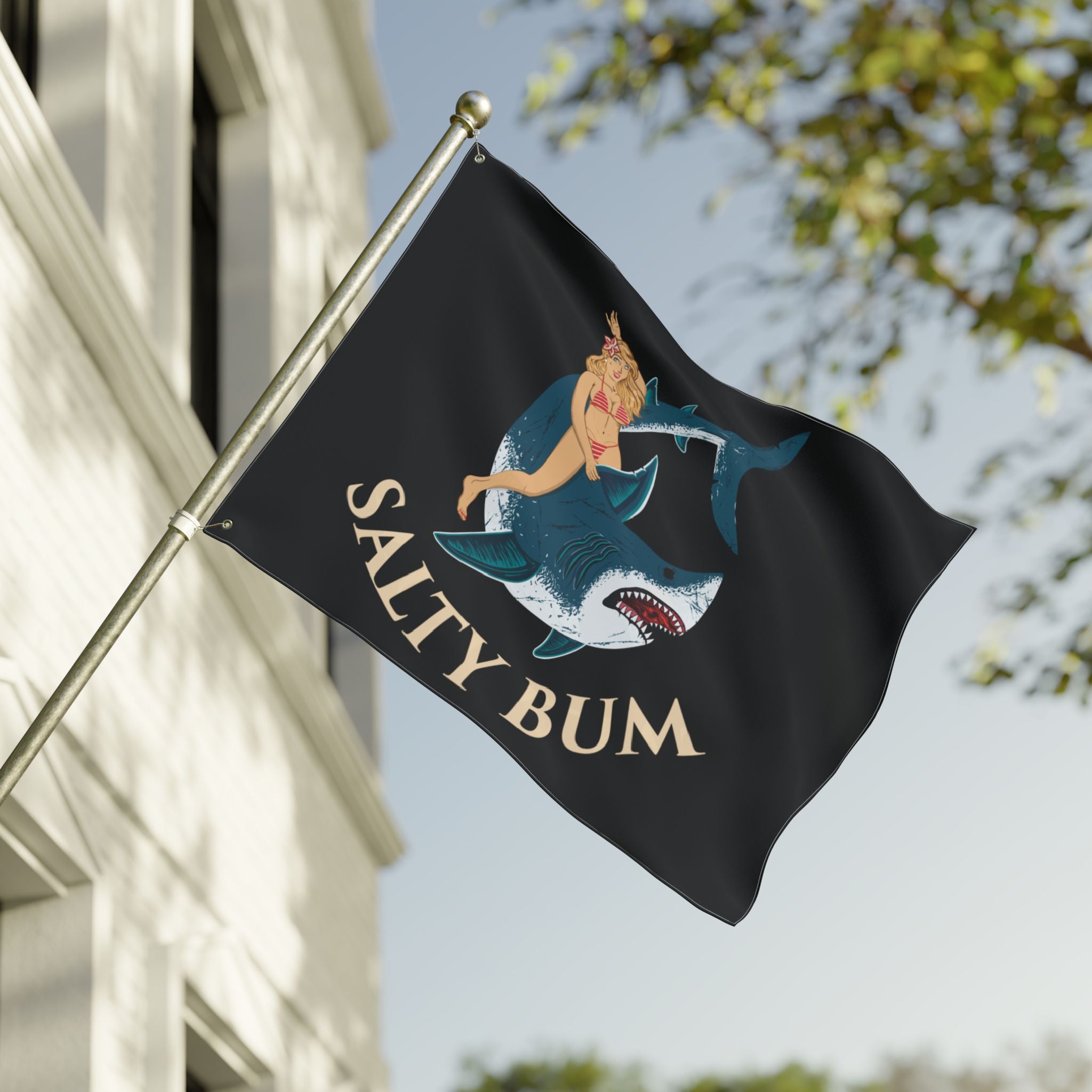 Flag with a shark and bikini-clad figure design and 'Salty Bum' text, waving in front of a building.