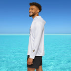 Man wearing a white long-sleeve shirt and black shorts standing on a beach with clear blue water and sky.