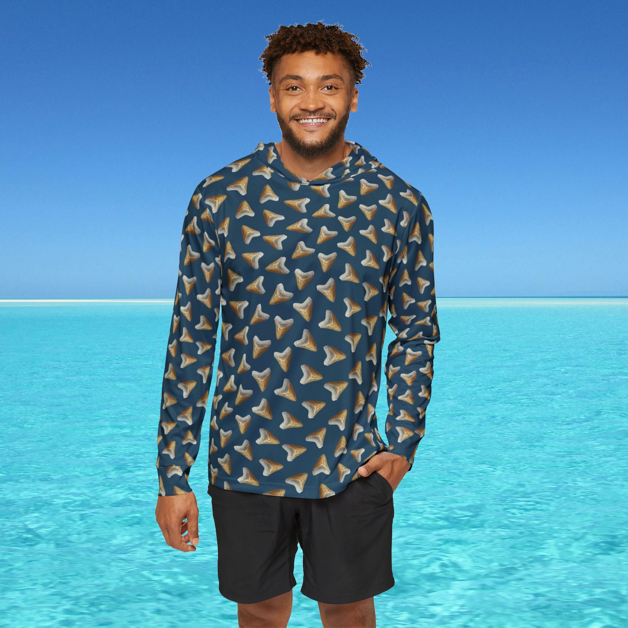 Man wearing a patterned long-sleeve shirt and black shorts standing in front of clear blue water and sky.