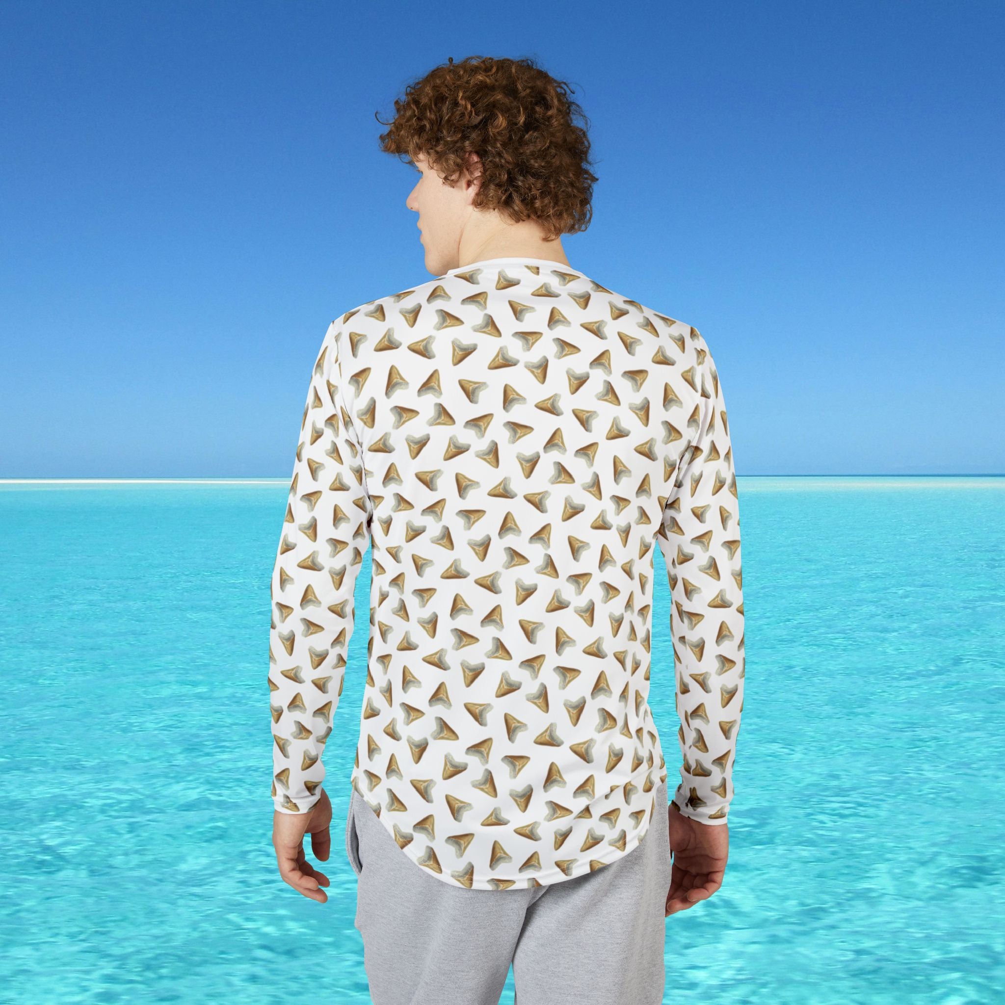 Man wearing a patterned shirt standing on a beach with clear blue water and sky.