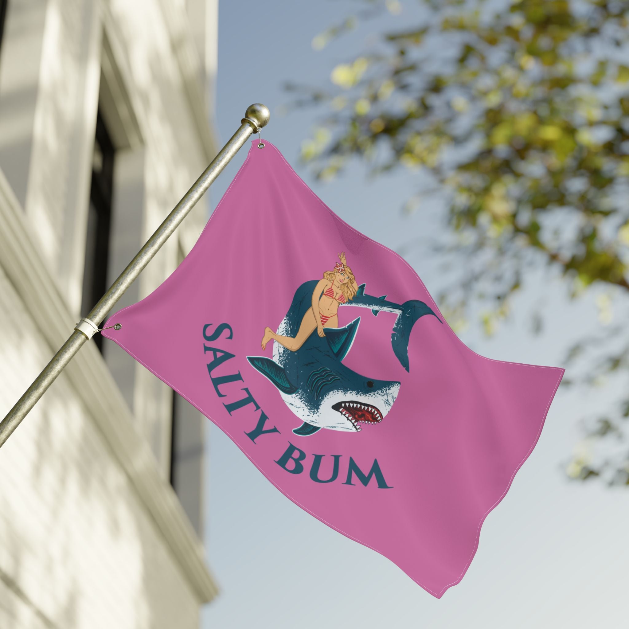 Pink flag with a mermaid and shark design and 'Salty Bum' text, waving in front of a building.
