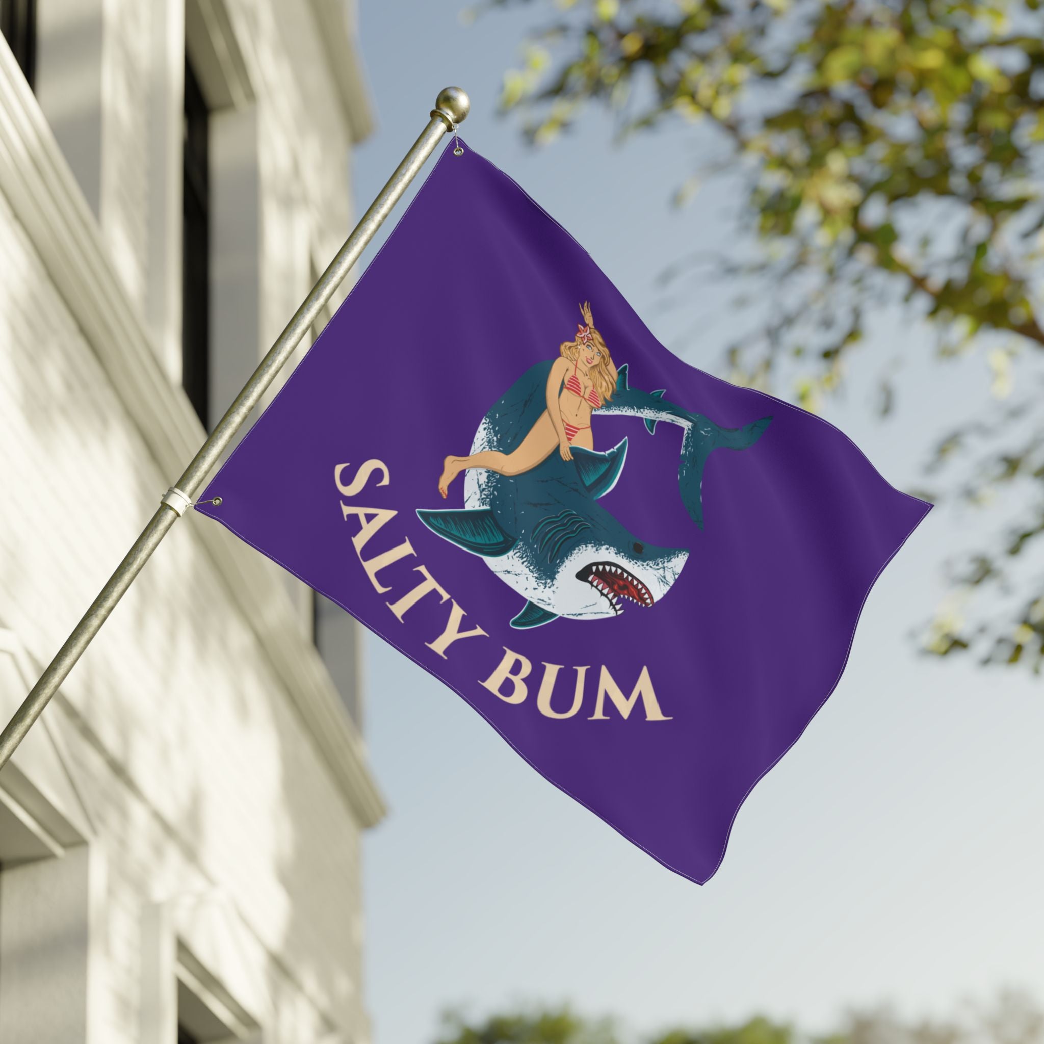 Flag with a mermaid and shark design on a purple background, featuring the text 'Salty Bum', waving in front of a building.