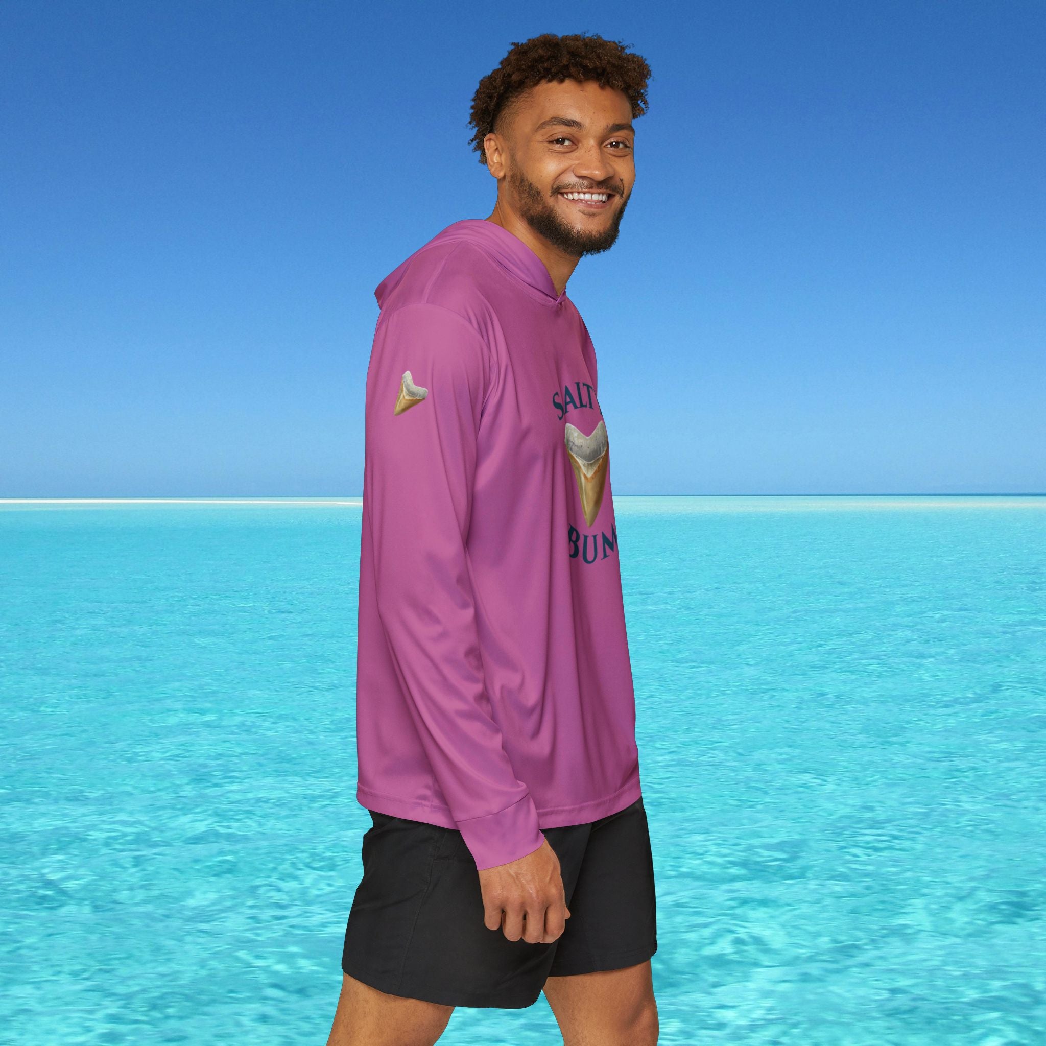 Man wearing a purple hoodie with text and graphics, standing in front of clear blue water and sky.