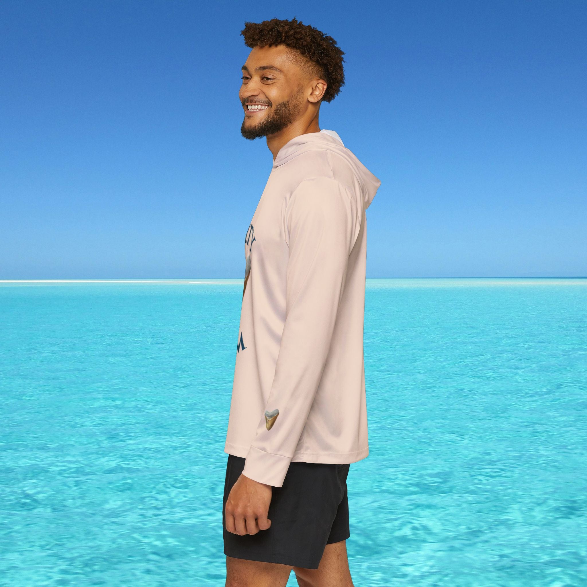 Man wearing a light pink hoodie and dark shorts standing on a beach with clear blue water and sky.