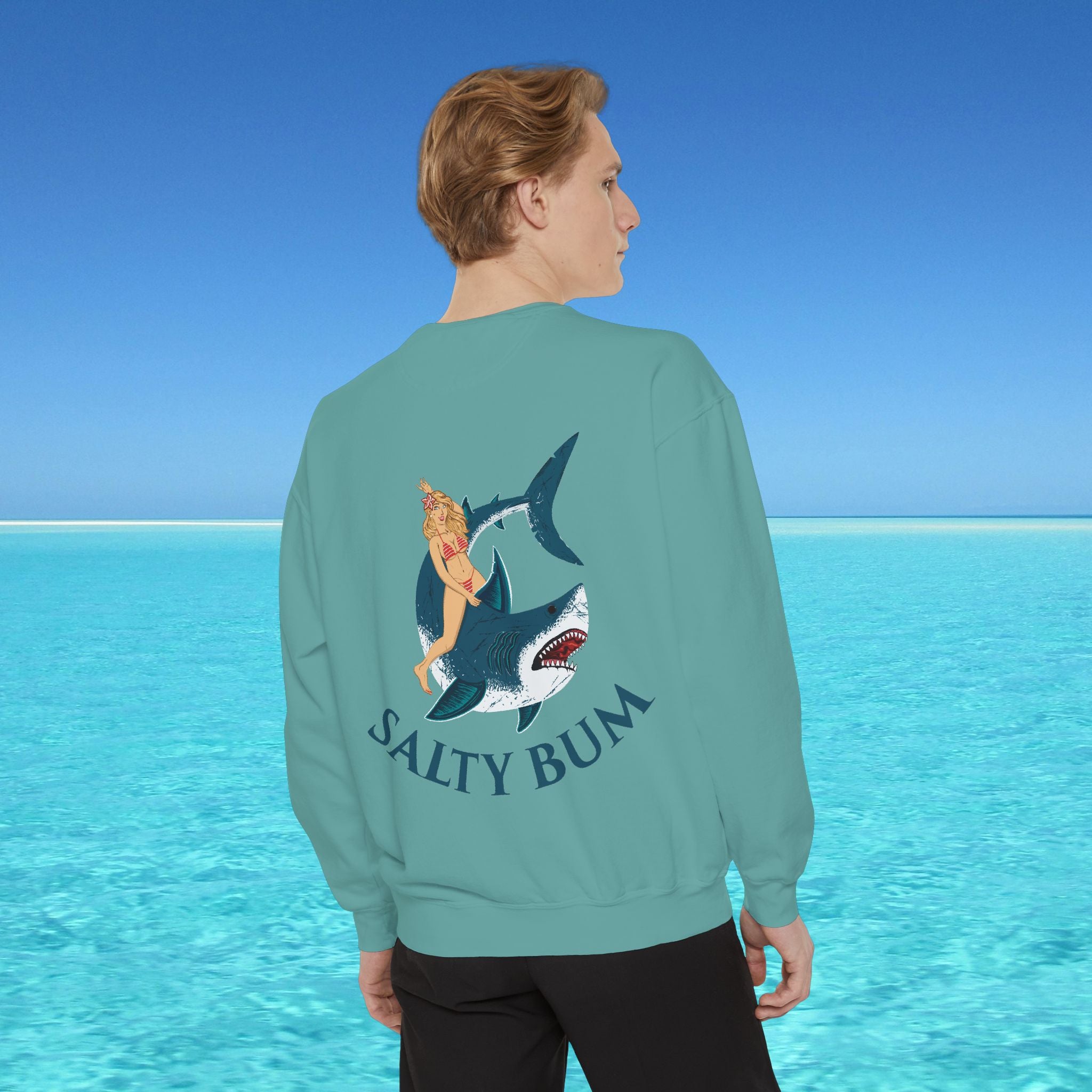 Person wearing a teal sweatshirt with a shark and bikini graphic and 'Salty Bum' text, standing in front of a blue ocean.
