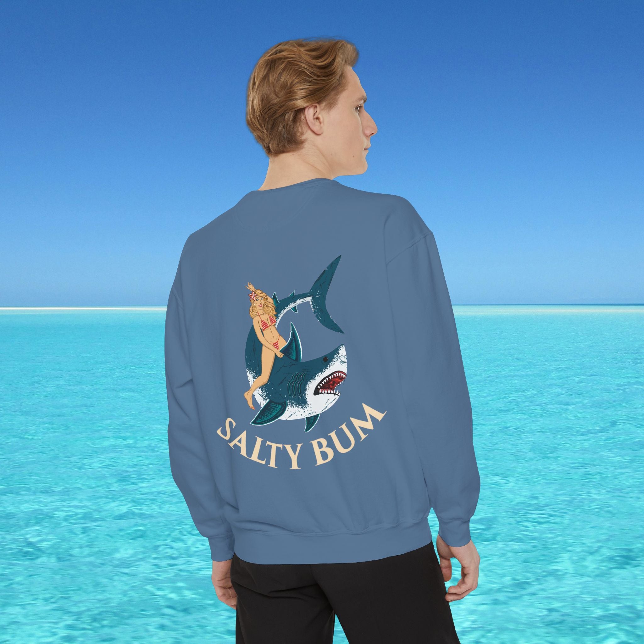 Person wearing a blue sweatshirt with a shark and bikini design, standing in front of a clear blue ocean.
