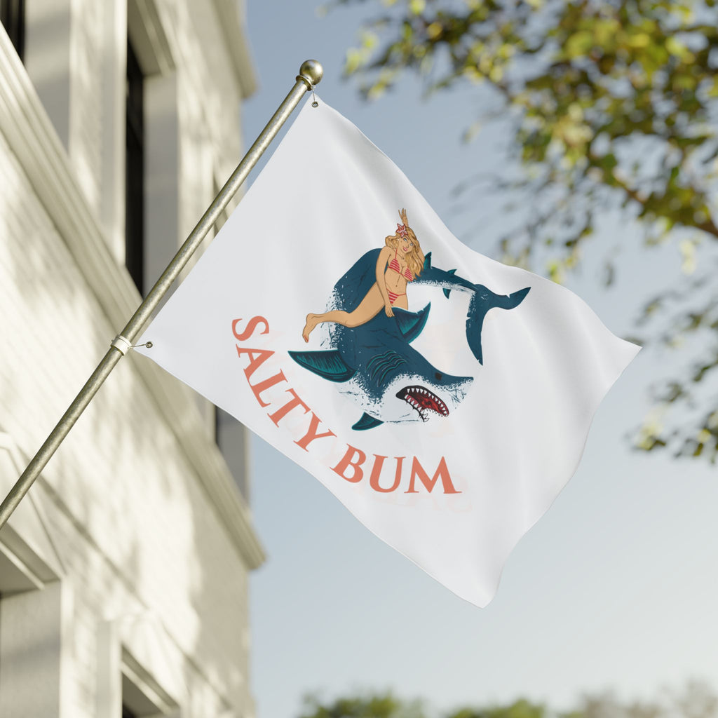 Flag with 'Salty Bum' design featuring a shark and person, waving in front of a building.