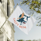 Flag with 'Salty Bum' design featuring a shark and person, waving in front of a building.