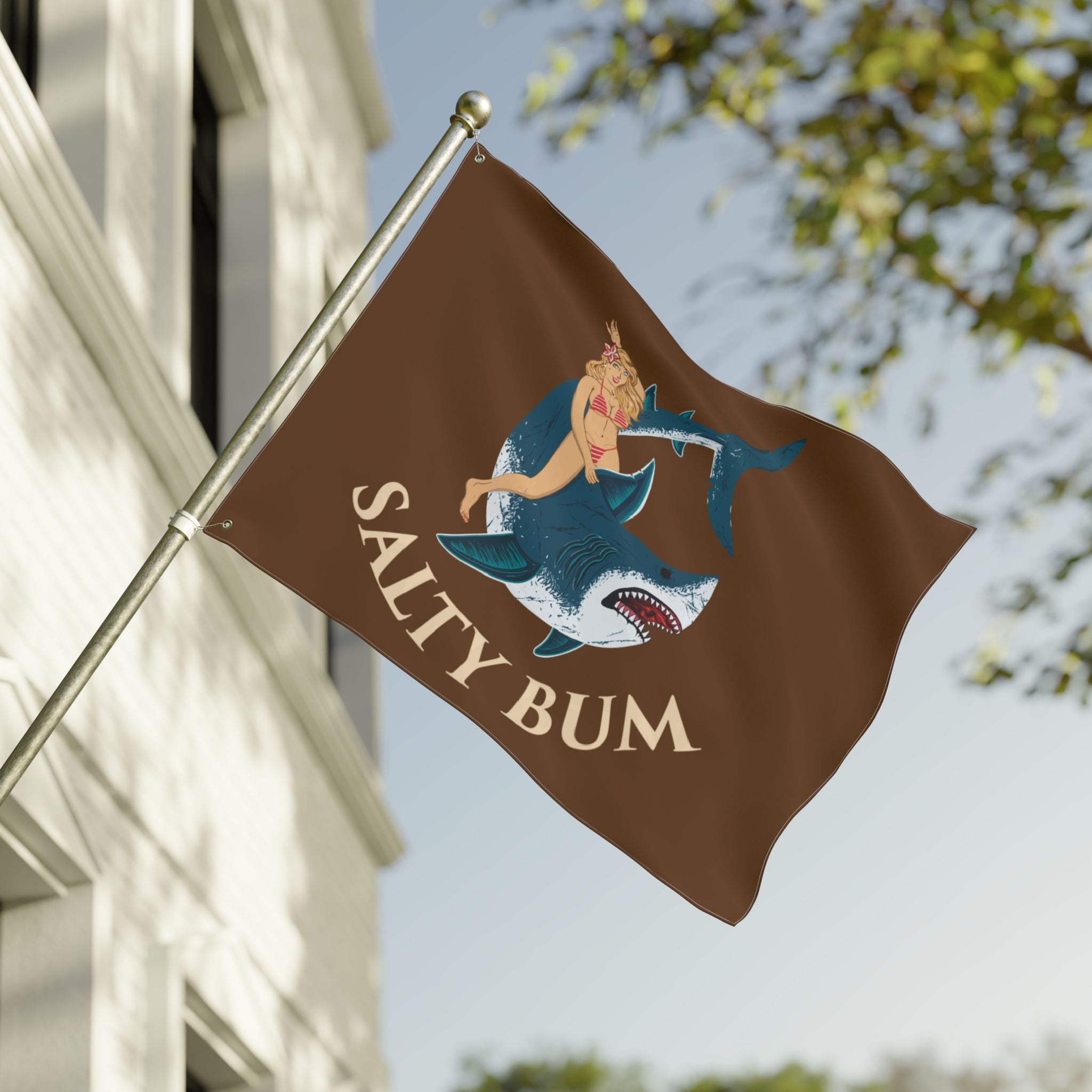 Flag with 'Salty Bum' design featuring a shark and a person, waving in front of a building.