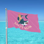Pink flag with a graphic design of a woman on a shark and 'Salty Bum' text, waving in front of a clear blue sky and ocean.