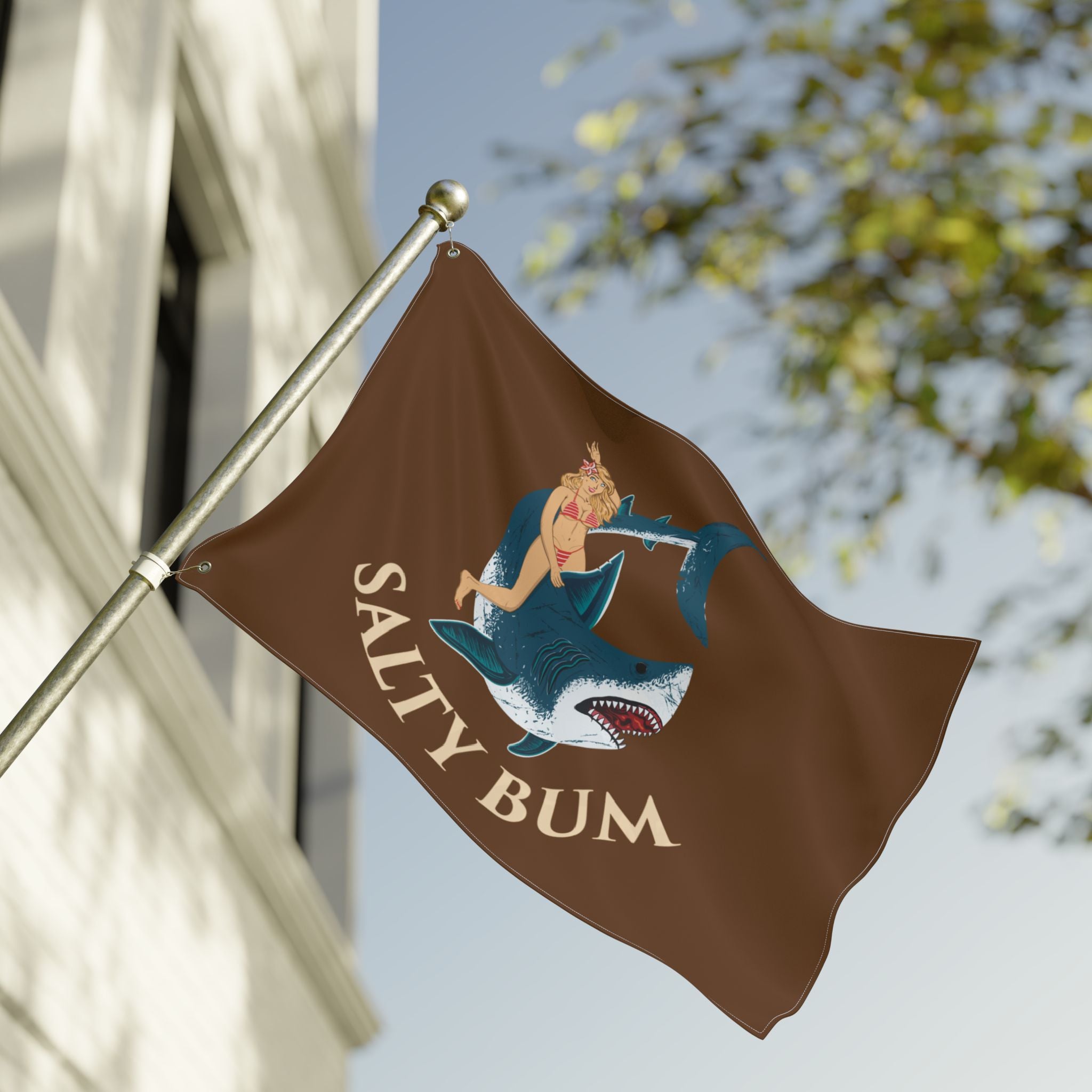 Flag with a graphic design of a woman on a shark and 'Salty Bum' text, waving in front of a building.