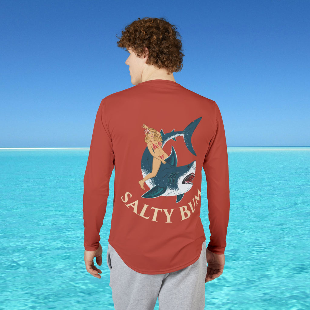 Person wearing a red long-sleeve shirt with a shark and mermaid graphic and 'Salty Bum' text, standing in front of clear blue water.