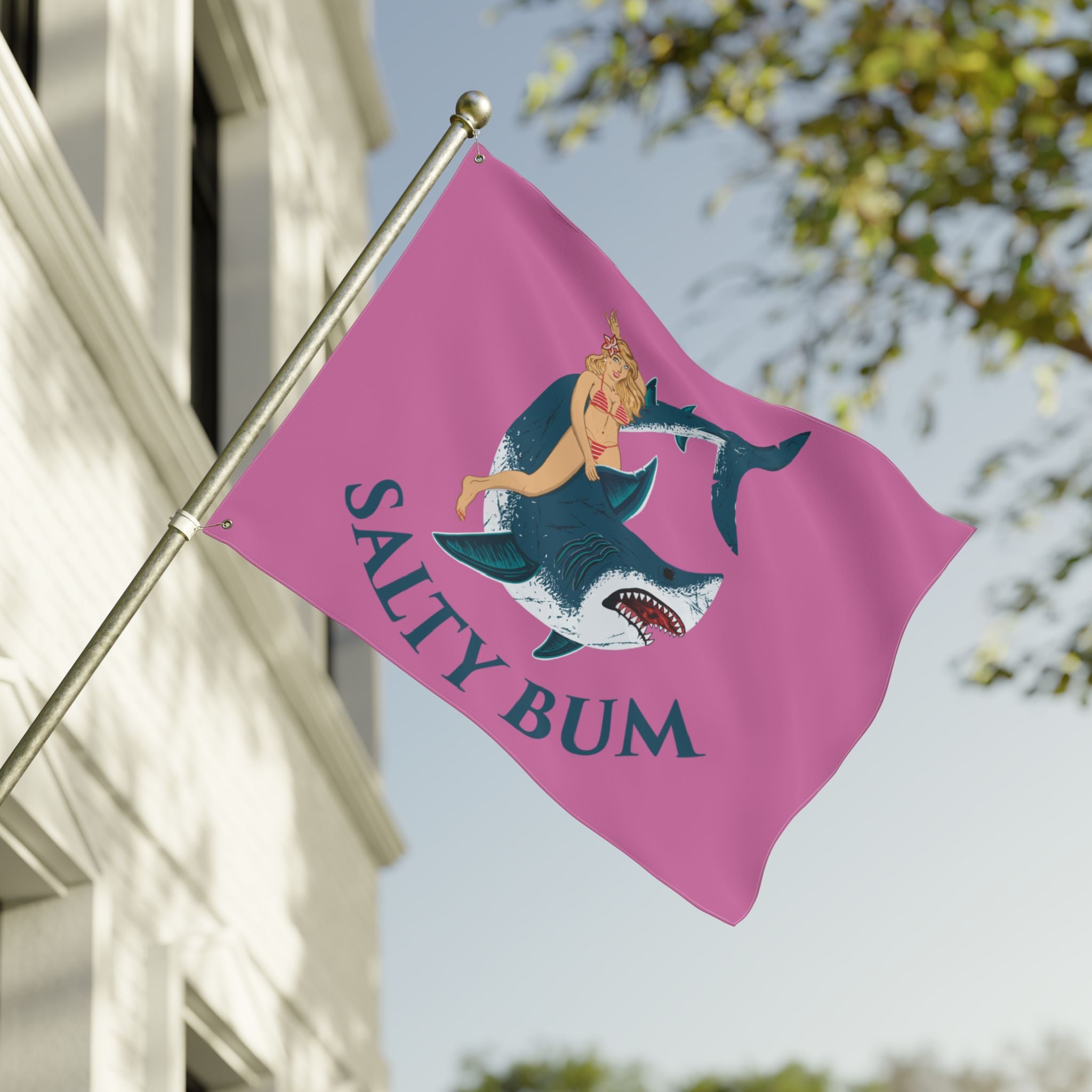 Pink flag with a mermaid and shark design and 'Salty Bum' text, waving in front of a building.
