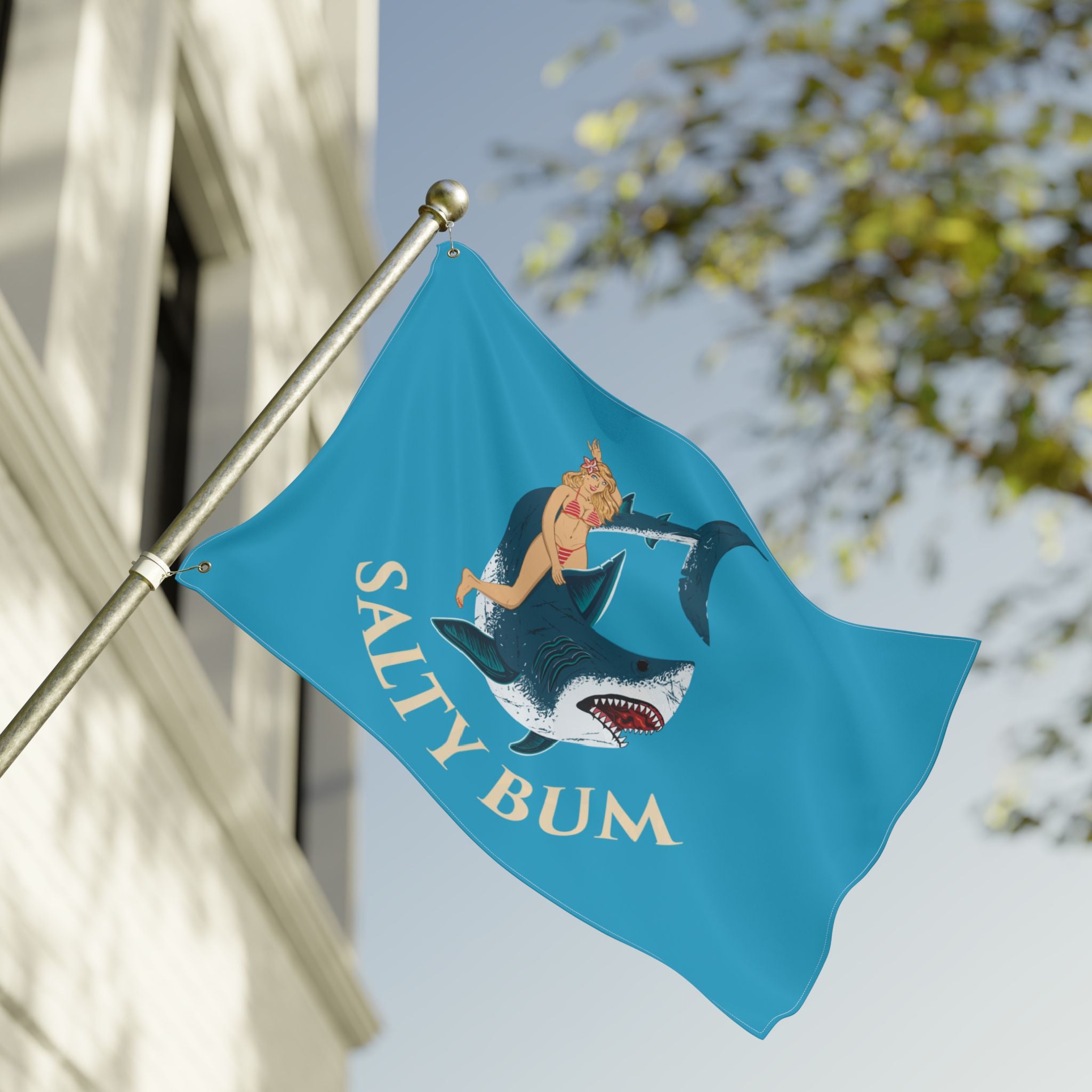 Flag with a shark and woman design and 'Salty Bum' text on a pole against a building and tree background.