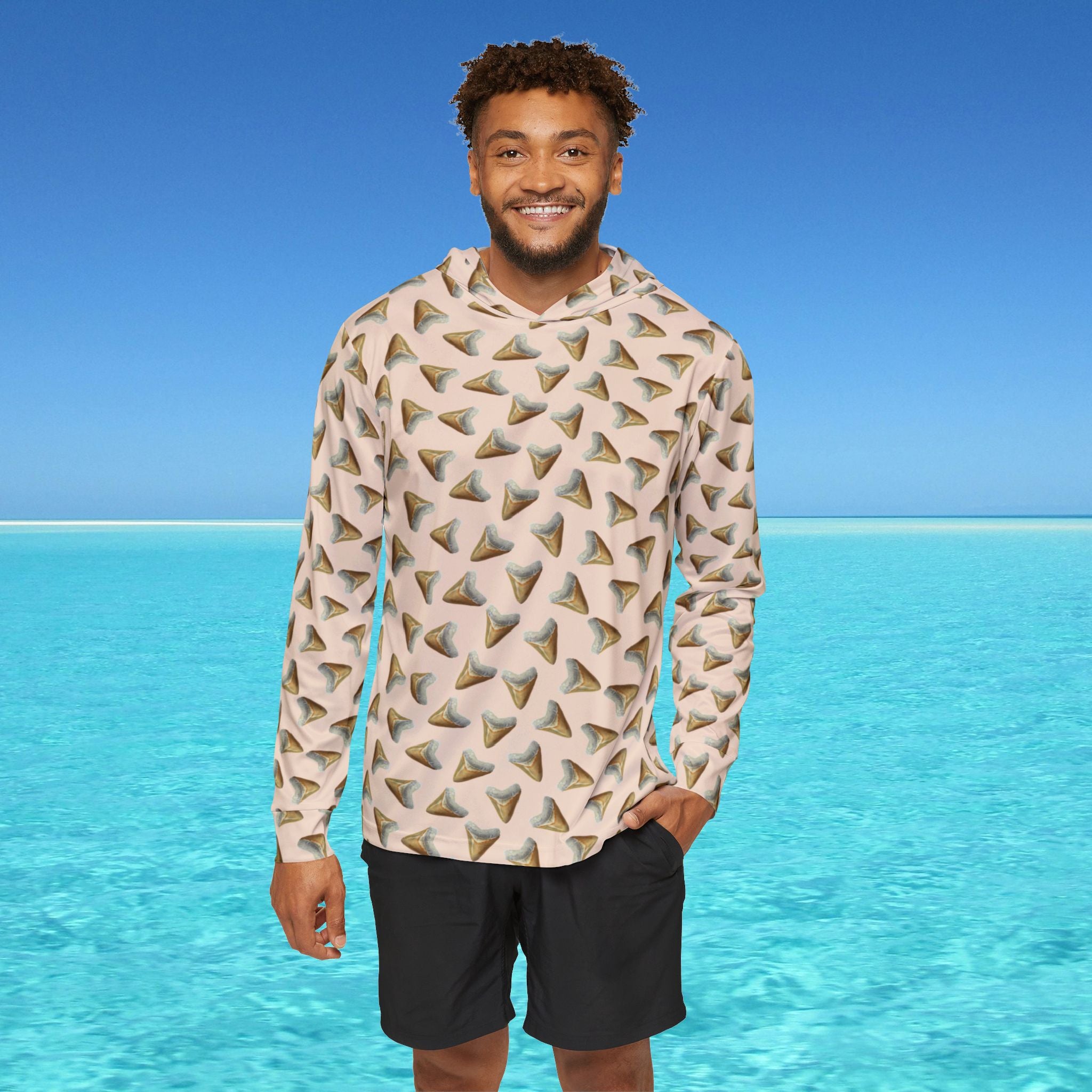 Man wearing a patterned hoodie and shorts standing in front of clear blue water and sky.