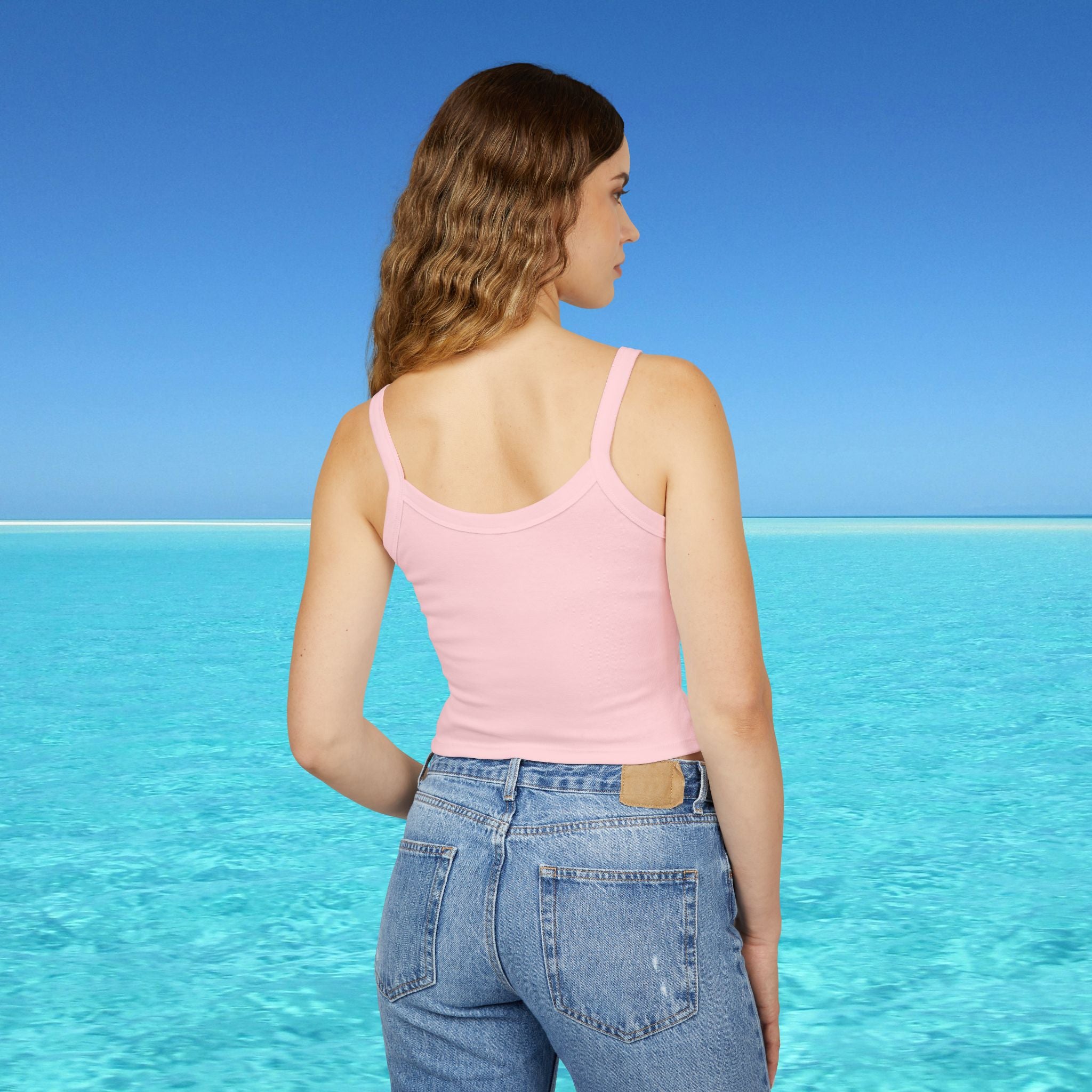 Woman wearing a pink tank top and blue jeans standing on a beach with clear blue water and sky.