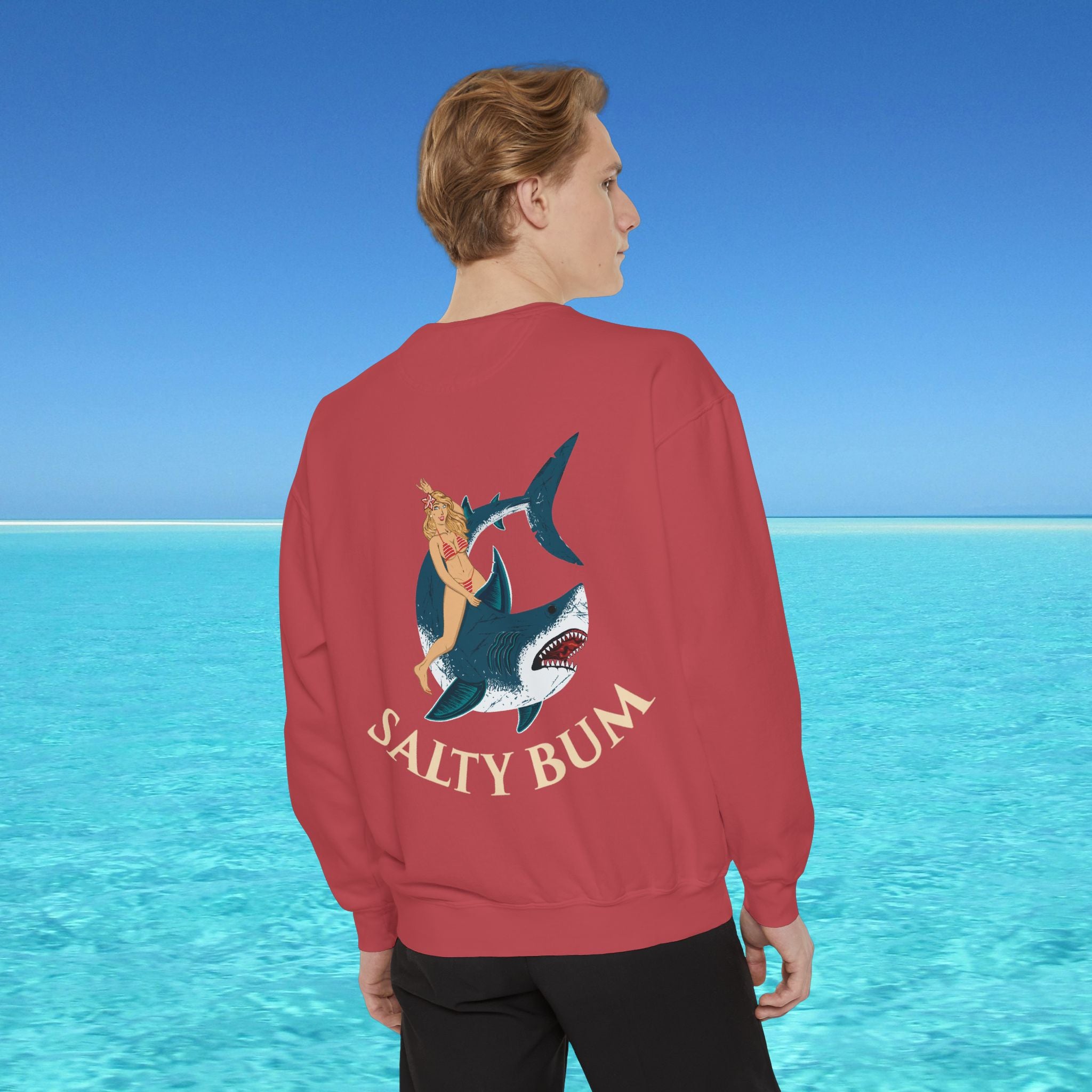 Person wearing a red sweatshirt with a shark and bikini design, standing in front of a blue ocean.