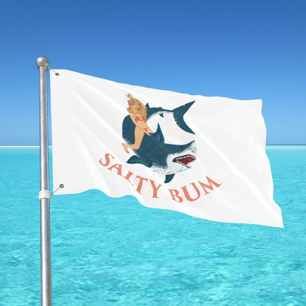 Flag with a shark and woman design and 'Salty Bum' text against a blue sky and ocean background