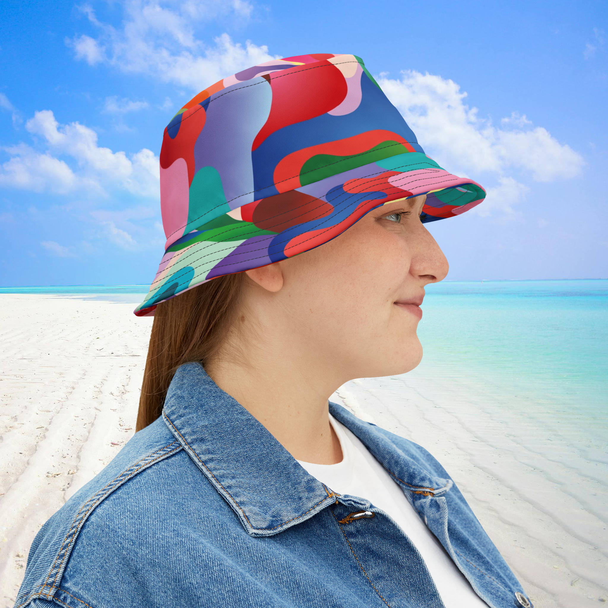 Person wearing a colorful bucket hat on a beach with blue sky and ocean.