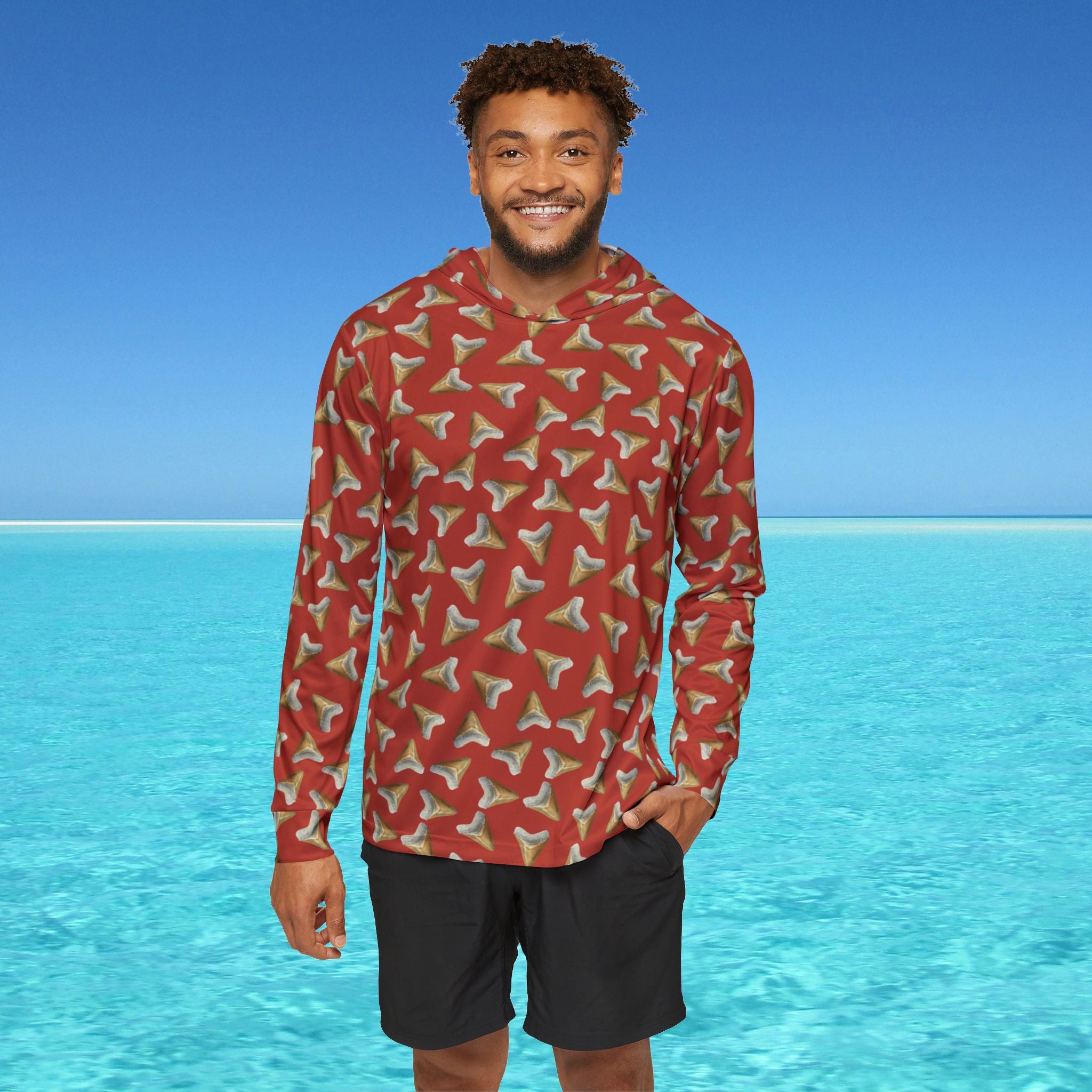 Man wearing a red patterned long-sleeve shirt and black shorts standing in front of clear blue water and sky.