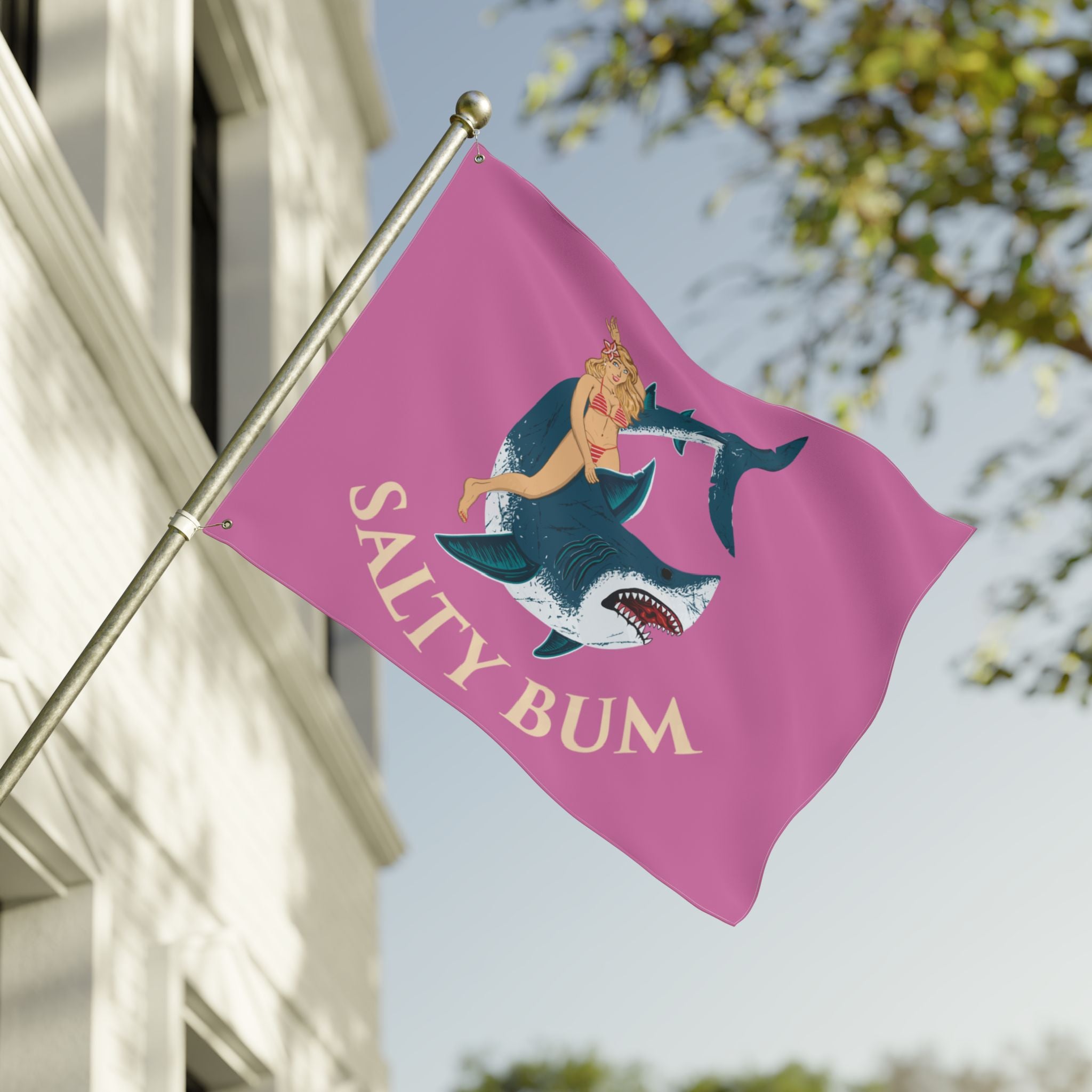 Flag with a shark and bikini-clad figure design on a pink background, featuring the text 'Salty Bum', waving in front of a building.