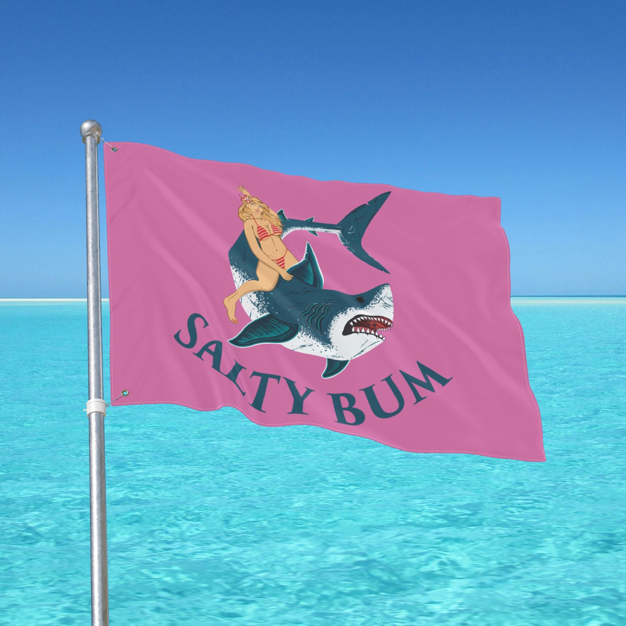 Flag with a shark and mermaid design on a pink background, waving in front of a clear blue sky and ocean.
