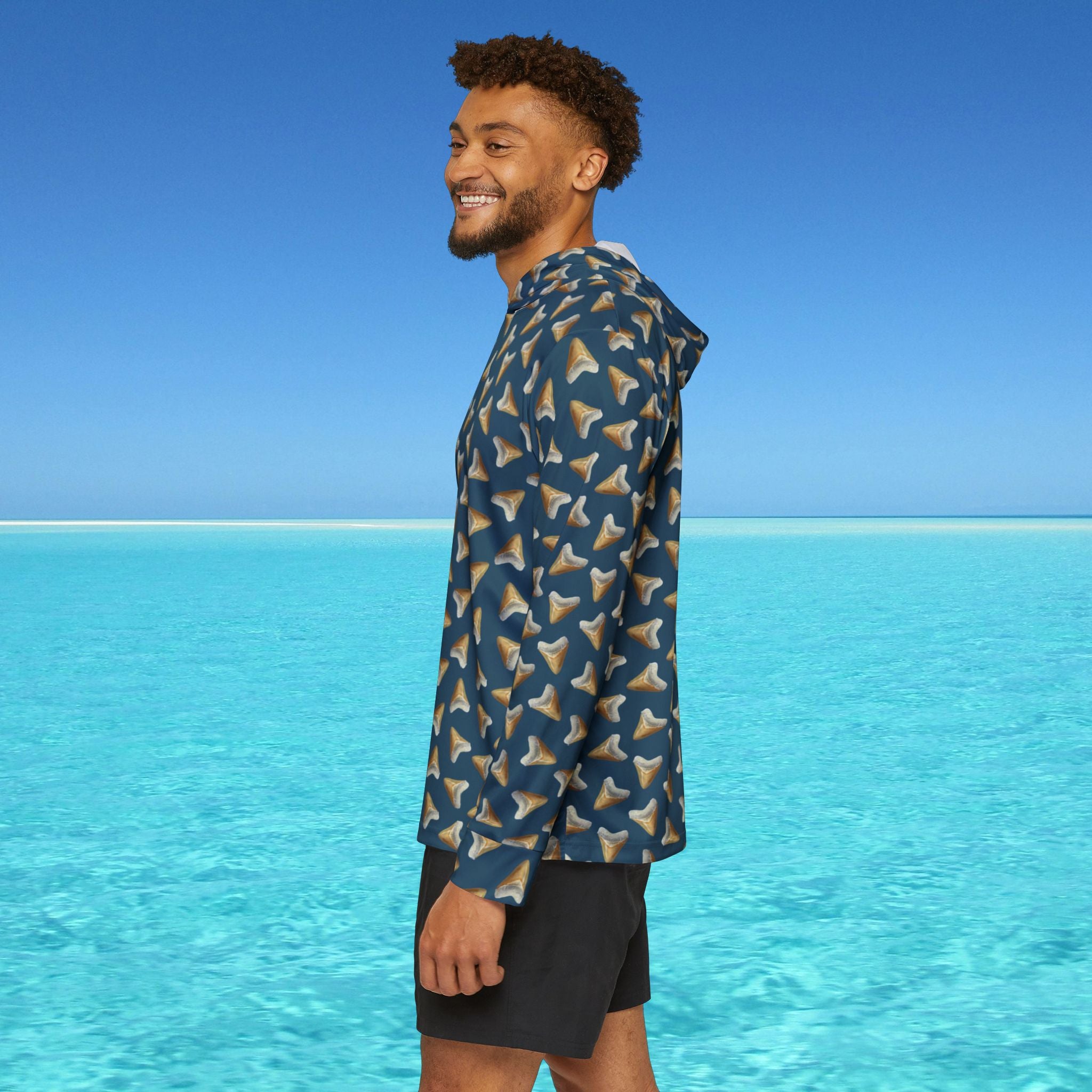 Man wearing a blue patterned shirt standing on a beach with clear blue water and sky.