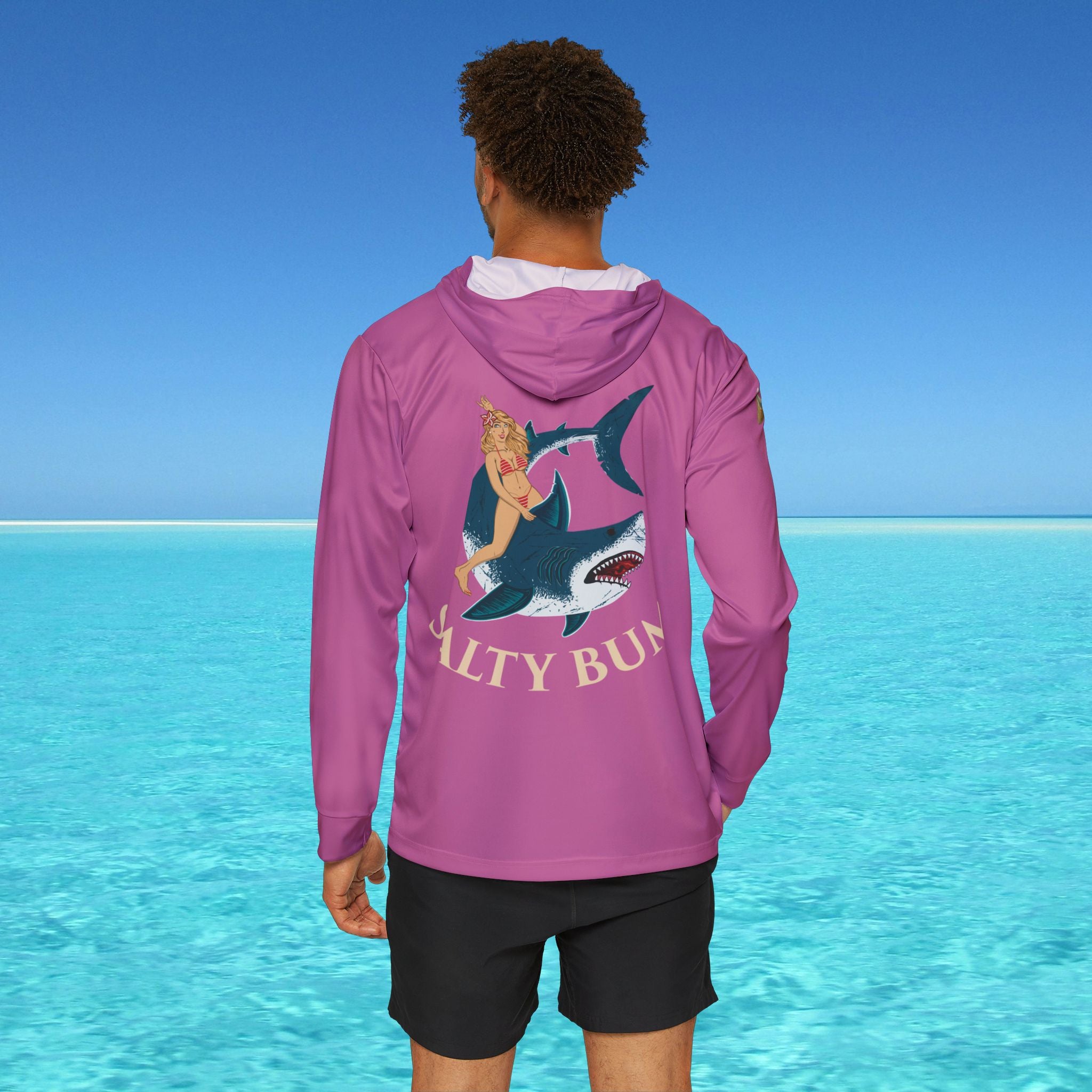 Person wearing a pink hoodie with a shark graphic and 'Salty Bun' text, standing in front of clear blue water.