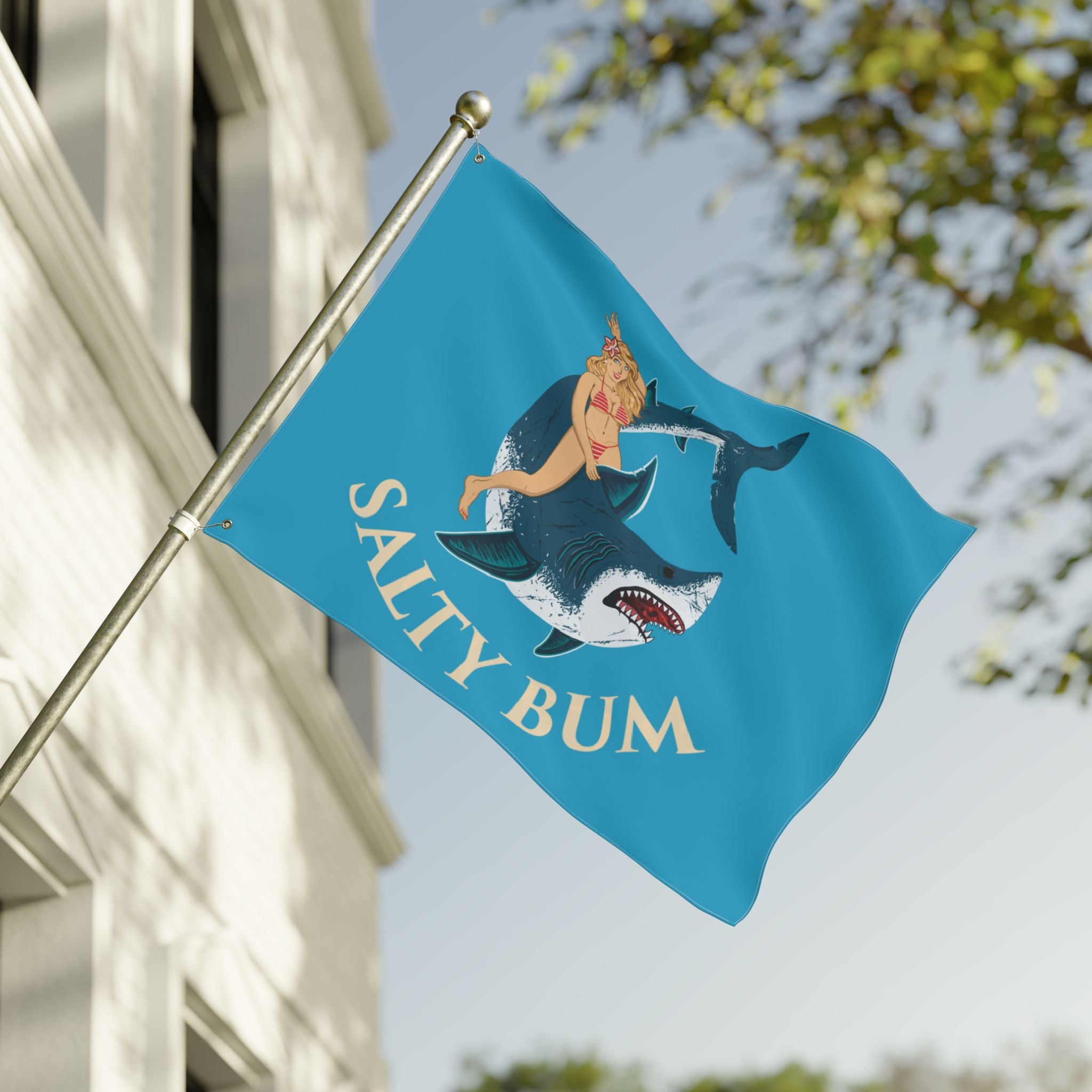 Flag with a shark and bikini-clad figure design on a blue background, featuring the text 'Salty Bum', waving in front of a building.