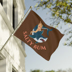 Flag with a shark and woman design and 'Salty Bum' text on a pole against a building and tree background.