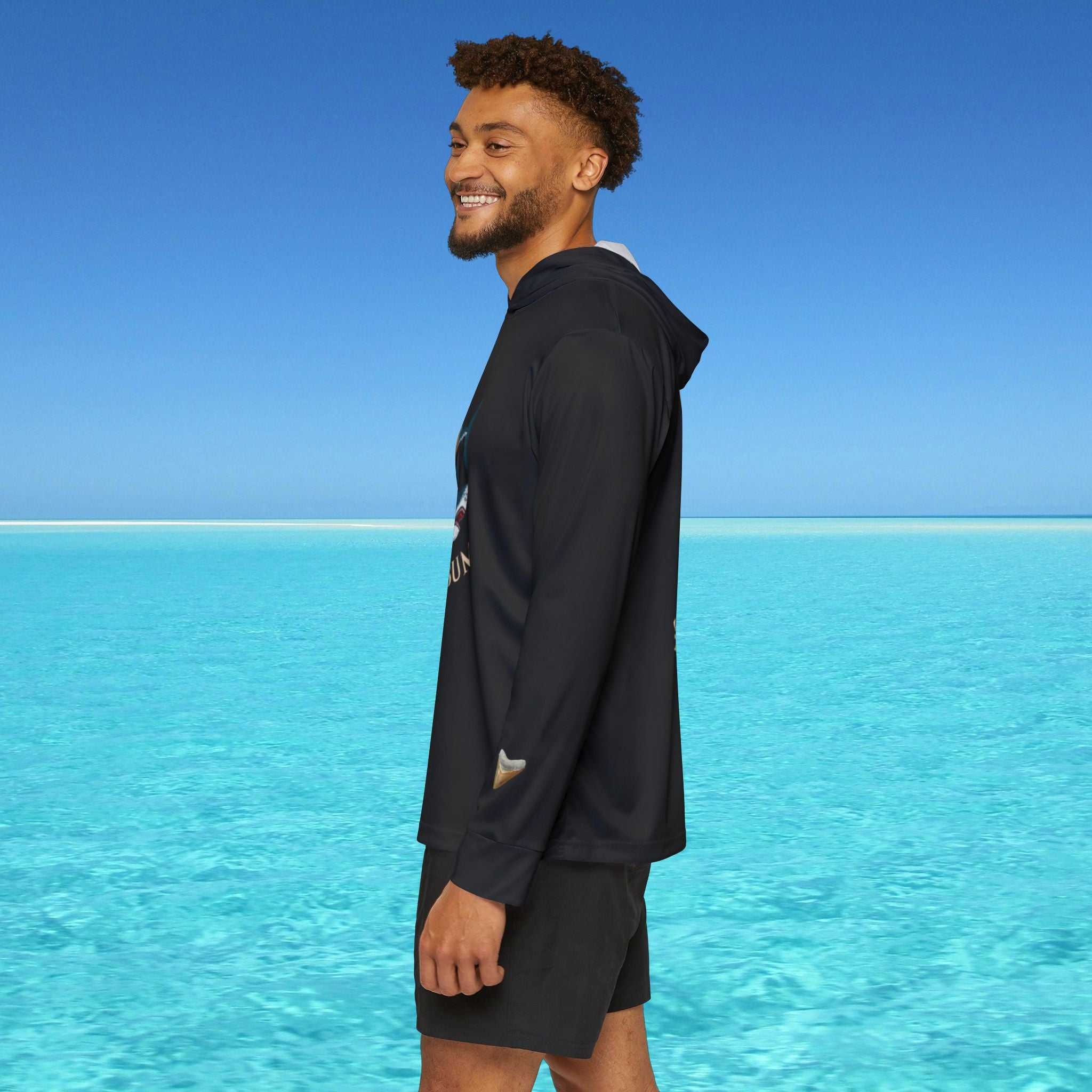 Man wearing a dark hoodie and shorts standing on a beach with clear blue water and sky.