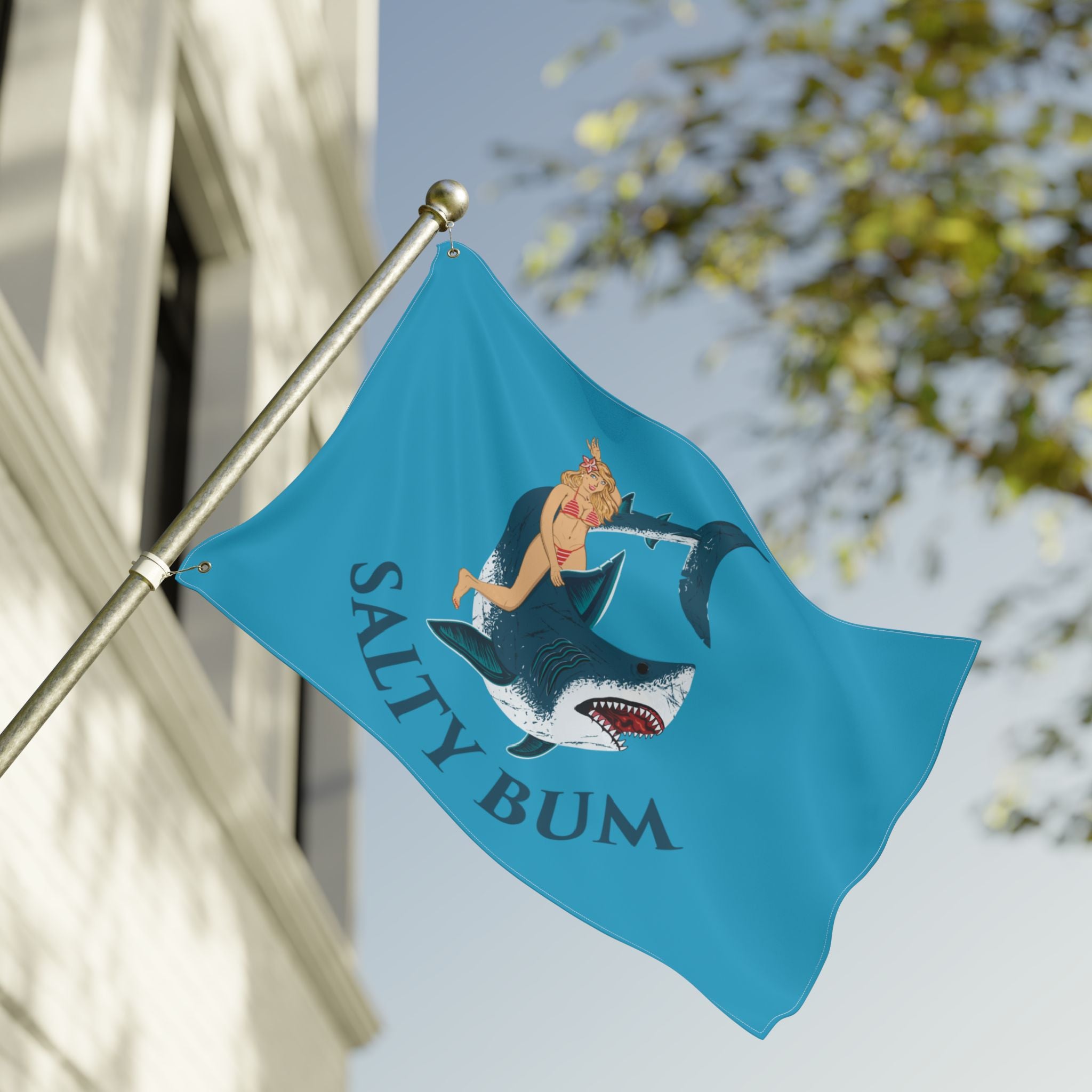 Blue flag with a shark and woman design and 'Salty Bum' text, waving in front of a building.