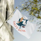 Flag with a graphic design of a woman on a shark and 'Salty Bum' text, waving outdoors.