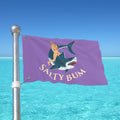 Flag with a shark and mermaid design on a pole against a blue sky and ocean.