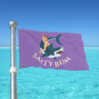 Flag with a shark and mermaid design on a pole against a blue sky and ocean.