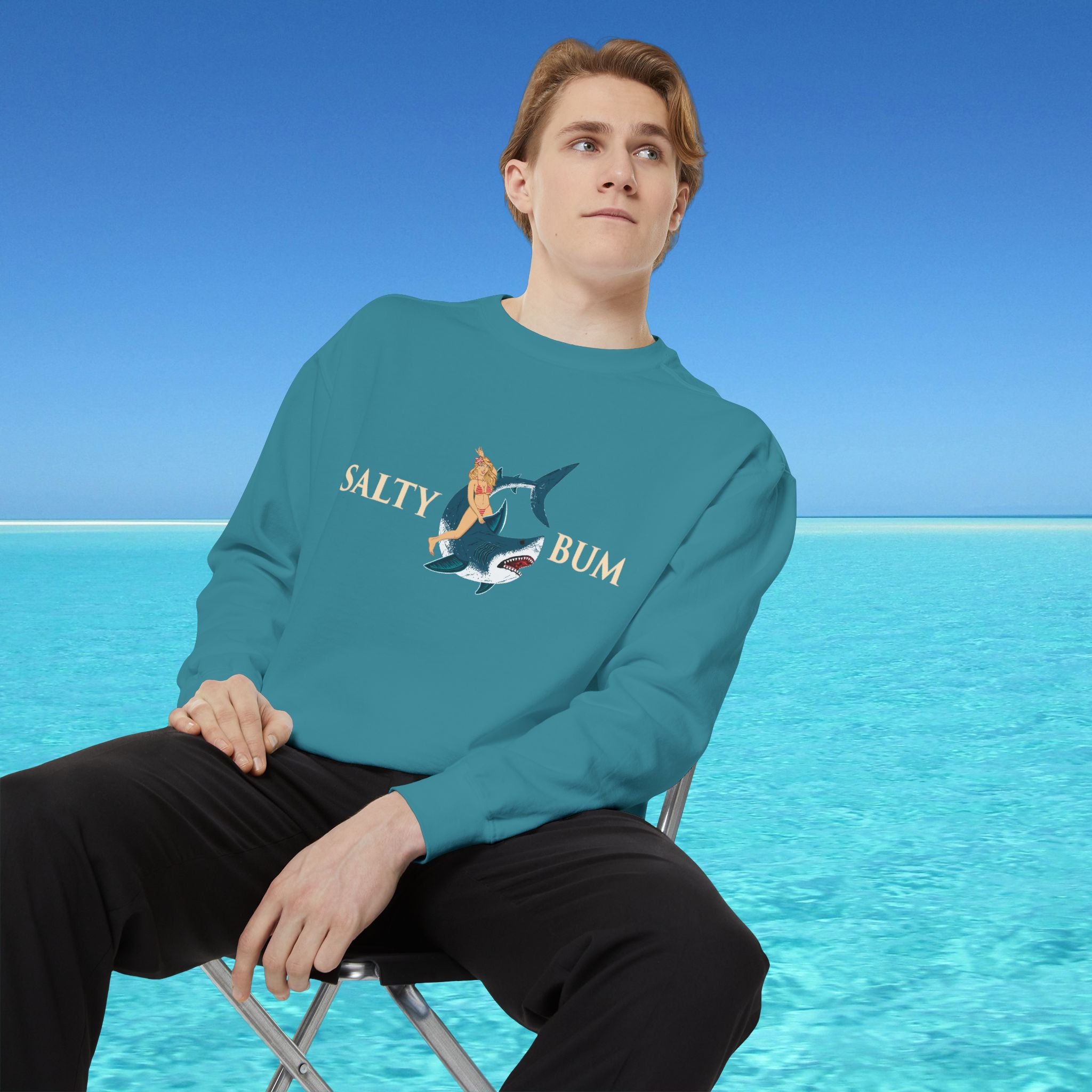 Person wearing a teal sweatshirt with a graphic design sitting on a chair in front of a blue ocean.
