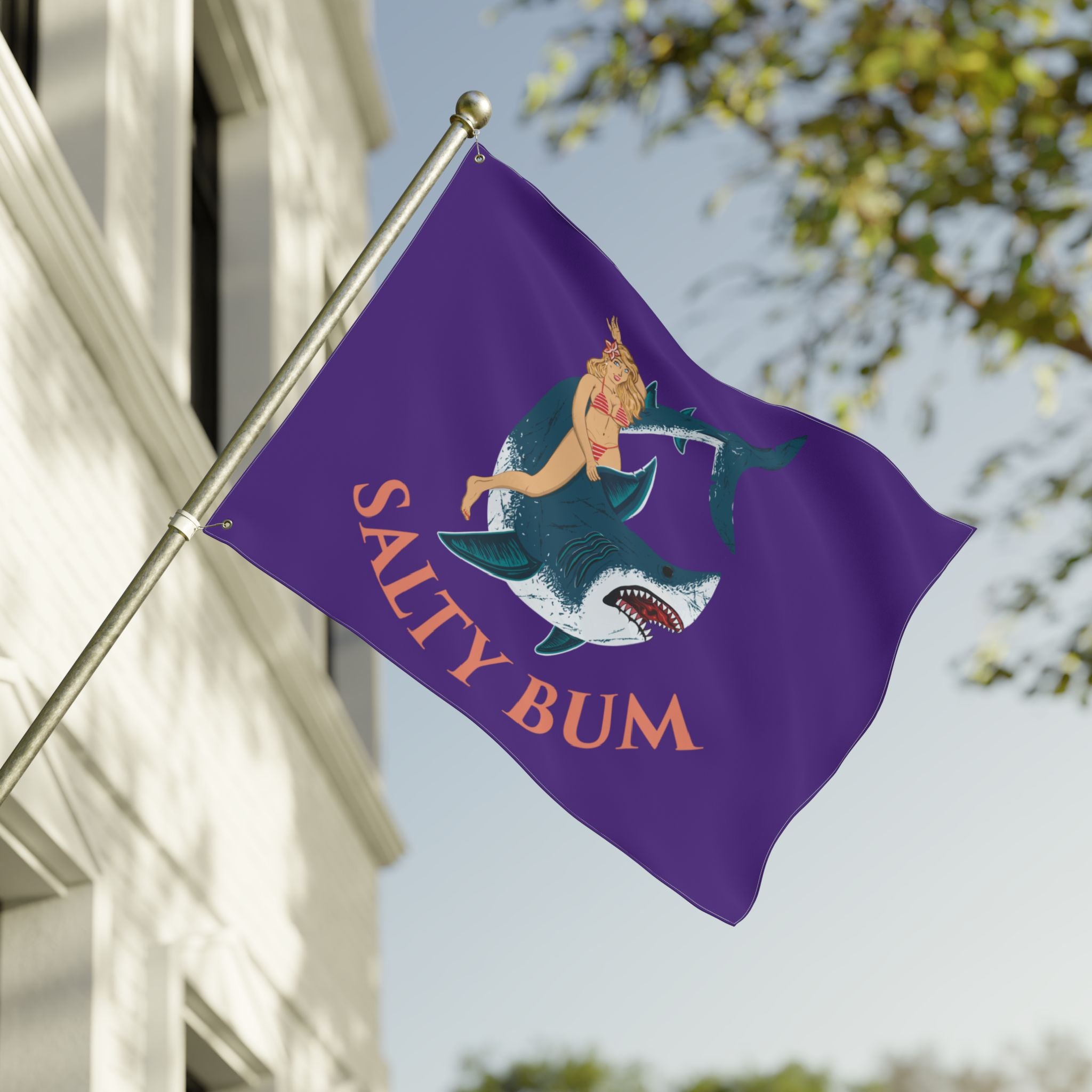 Purple flag with a shark and mermaid design and 'Salty Bum' text, waving in front of a building.