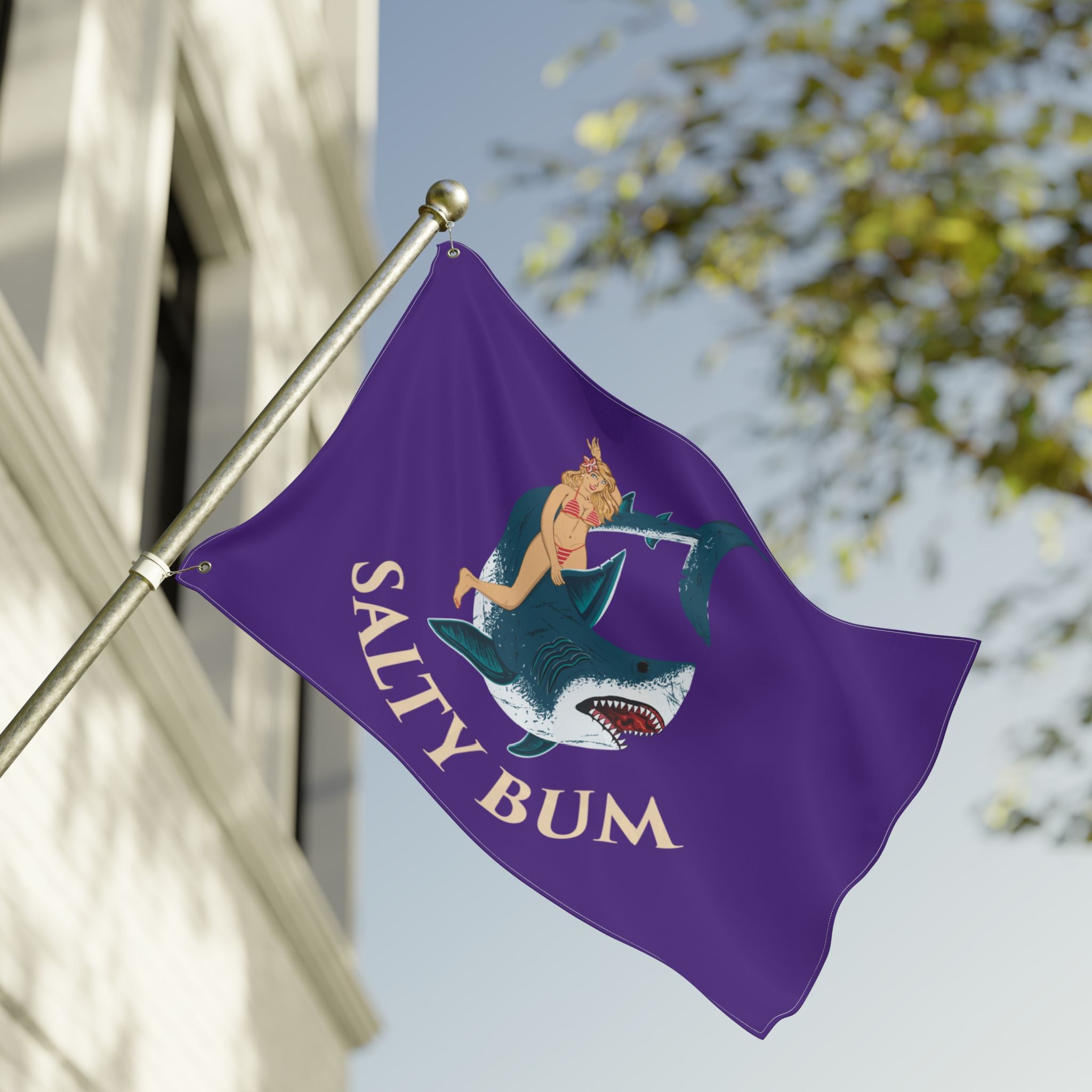 Purple flag with a mermaid and shark design, featuring the text 'Salty Bum', waving in front of a building.