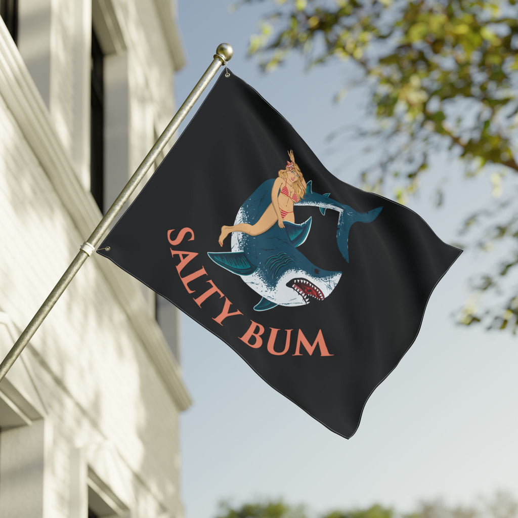 Flag with a shark and woman design and 'Salty Bum' text on a pole against a building and tree background.