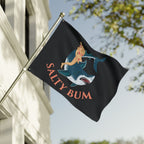 Flag with a shark and woman design and 'Salty Bum' text on a pole against a building and tree background.