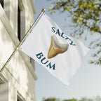 Flag with 'Salty Bum' text and a shark fin graphic waving in front of a building.