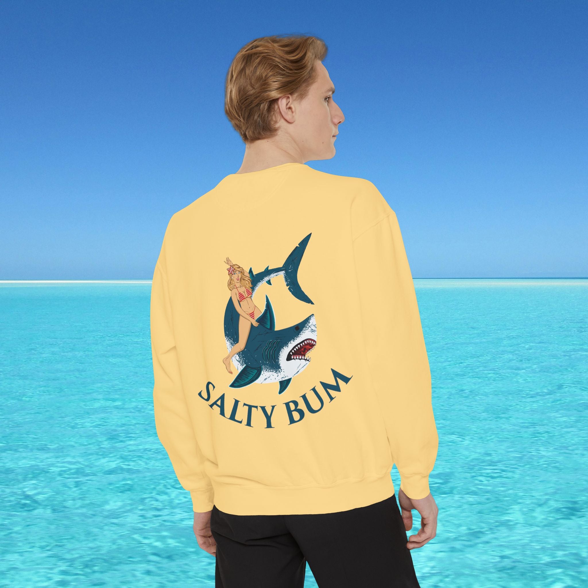 Person wearing a yellow sweatshirt with a shark graphic and 'Salty Bum' text, standing in front of a blue ocean.