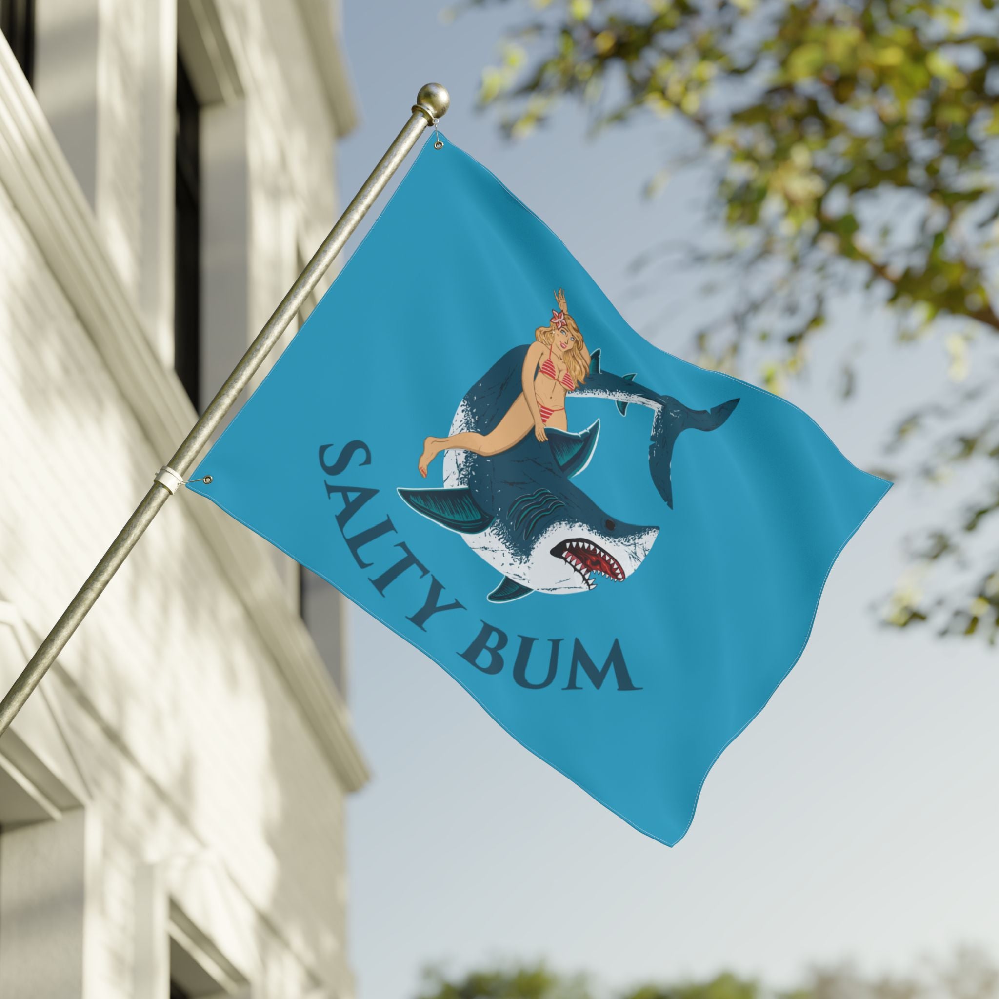 Flag with a shark and bikini-clad figure design on a blue background, featuring the text 'Salty Bum', waving in front of a building.
