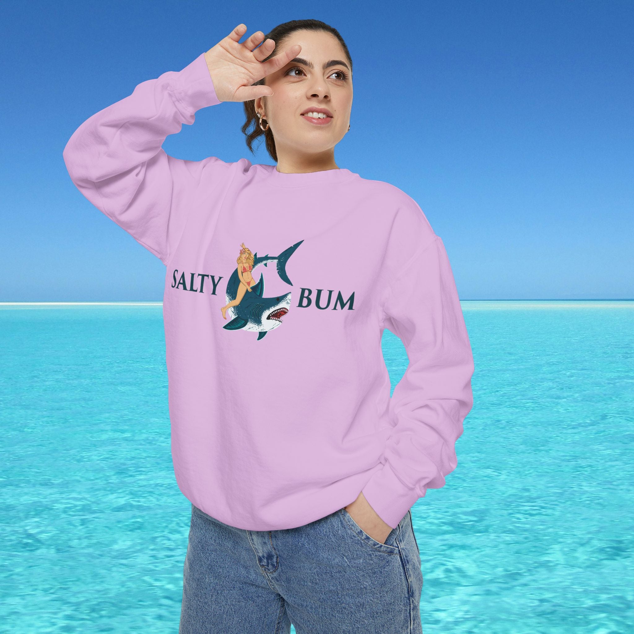 Person wearing a pink sweatshirt with a graphic design and text, standing against a blue sky and water background.