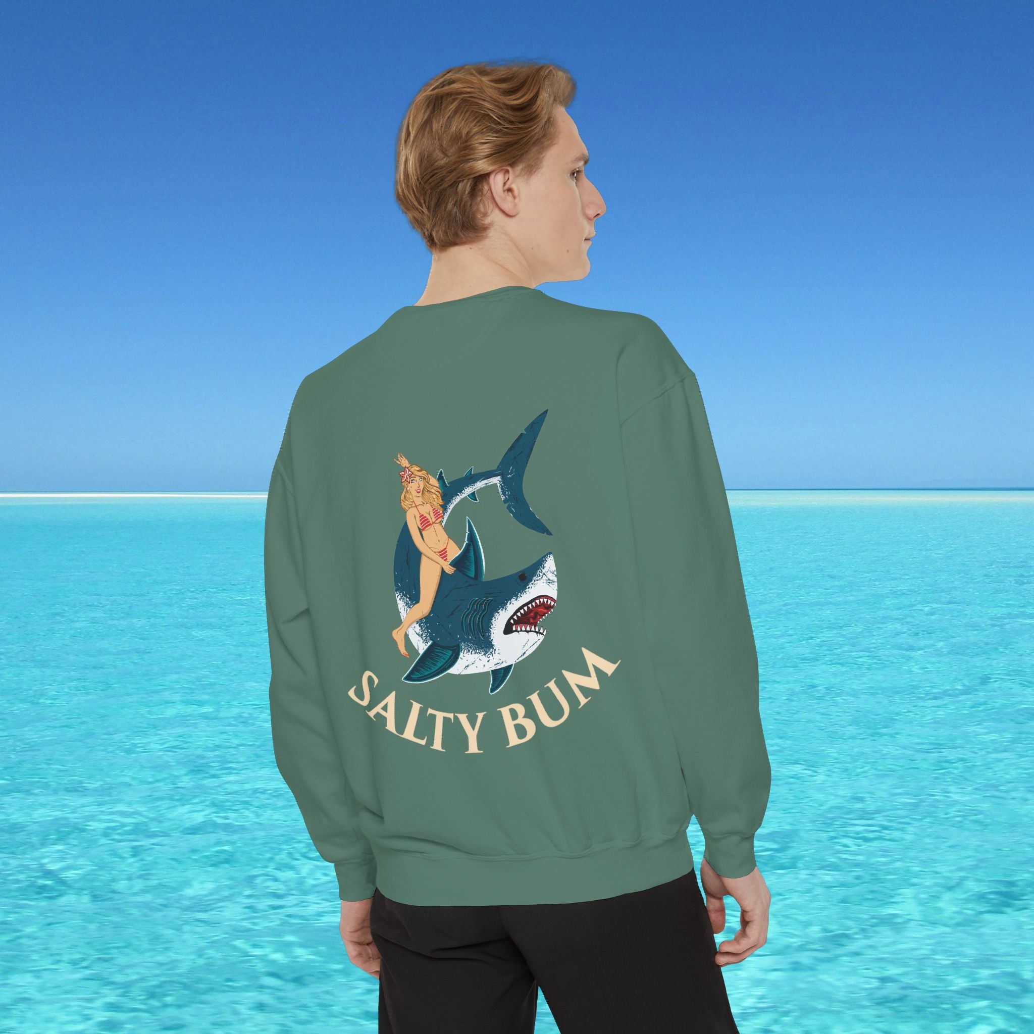 Person wearing a green sweatshirt with a shark and bikini graphic and 'Salty Bum' text, standing in front of a blue ocean.