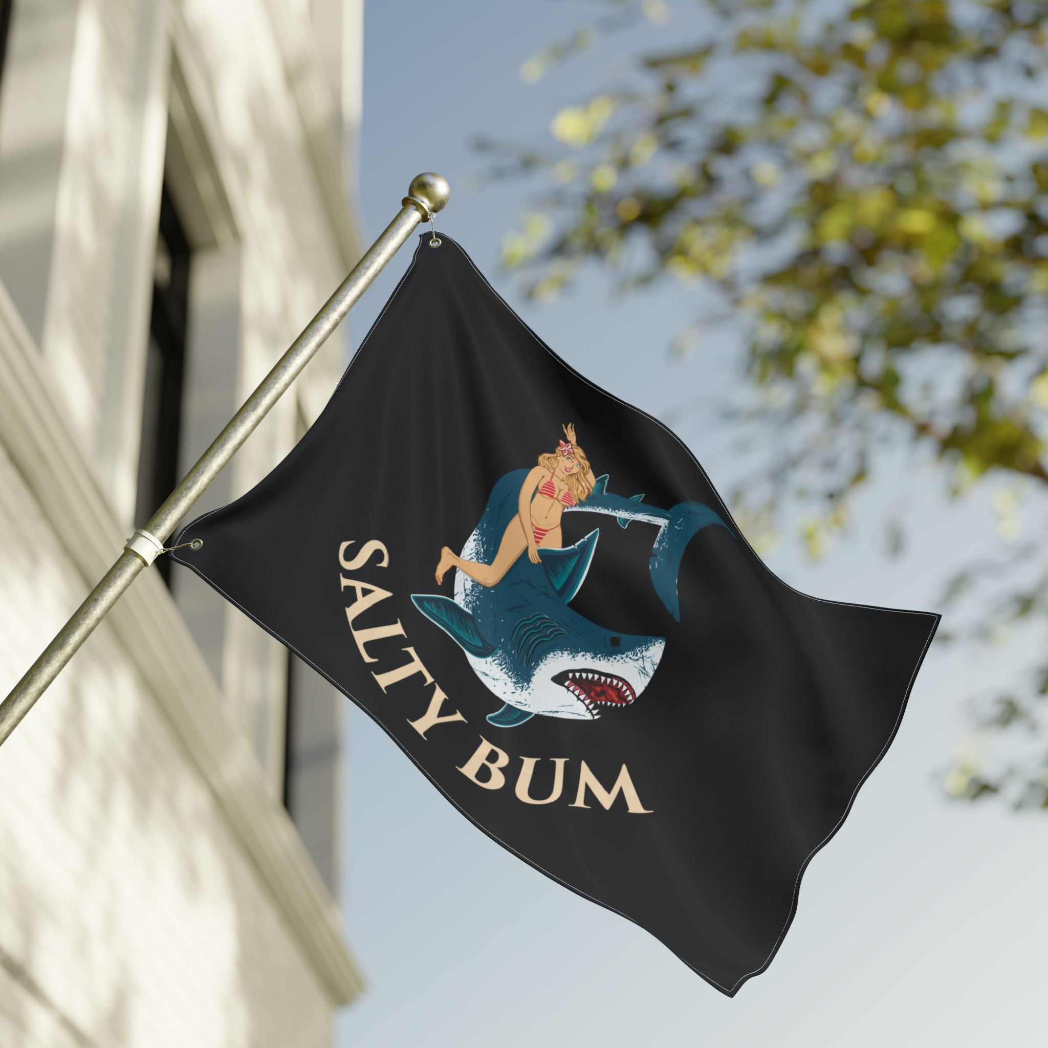 Flag with a graphic design of a woman on a shark and 'Salty Bum' text, waving in front of a building.