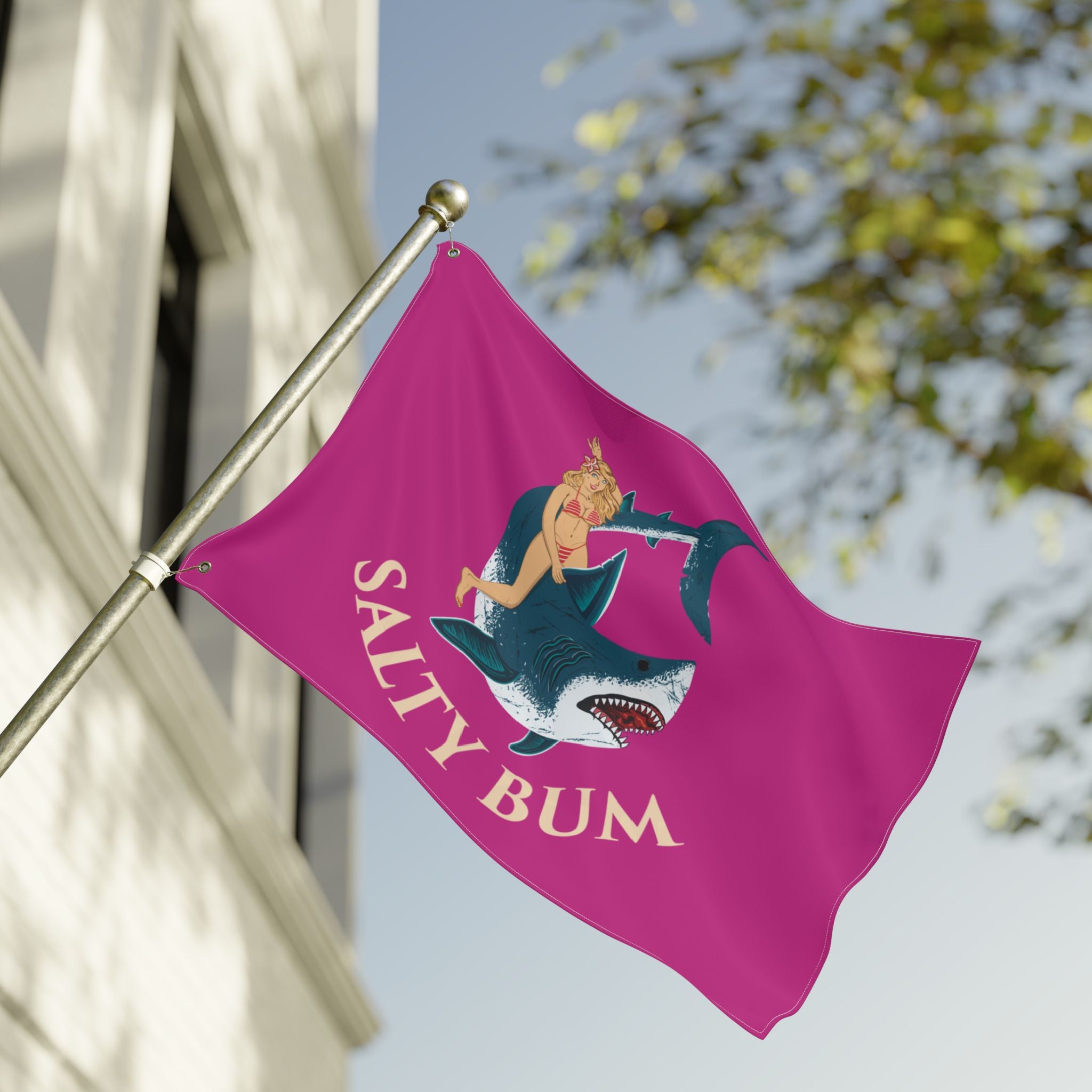 Pink flag with a graphic of a woman on a shark and 'Salty Bum' text, attached to a flagpole outdoors.